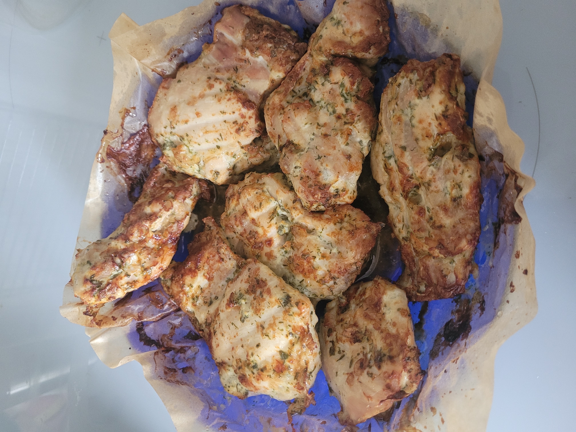 The image shows a pile of cooked chicken pieces on a piece of parchment paper. The chicken pieces are golden brown and appear to be seasoned with herbs. The parchment paper is slightly crinkled and has some dark brown spots, suggesting it was used for baking. The chicken is arranged in a shallow dish or on a tray, with a glimpse of a blue surface underneath the parchment paper. The lighting suggests the photo was taken indoors, likely in a kitchen environment. There are no people visible in the image. There is no information in the image to suggest this scene is located in Jūrmala, Latvia.