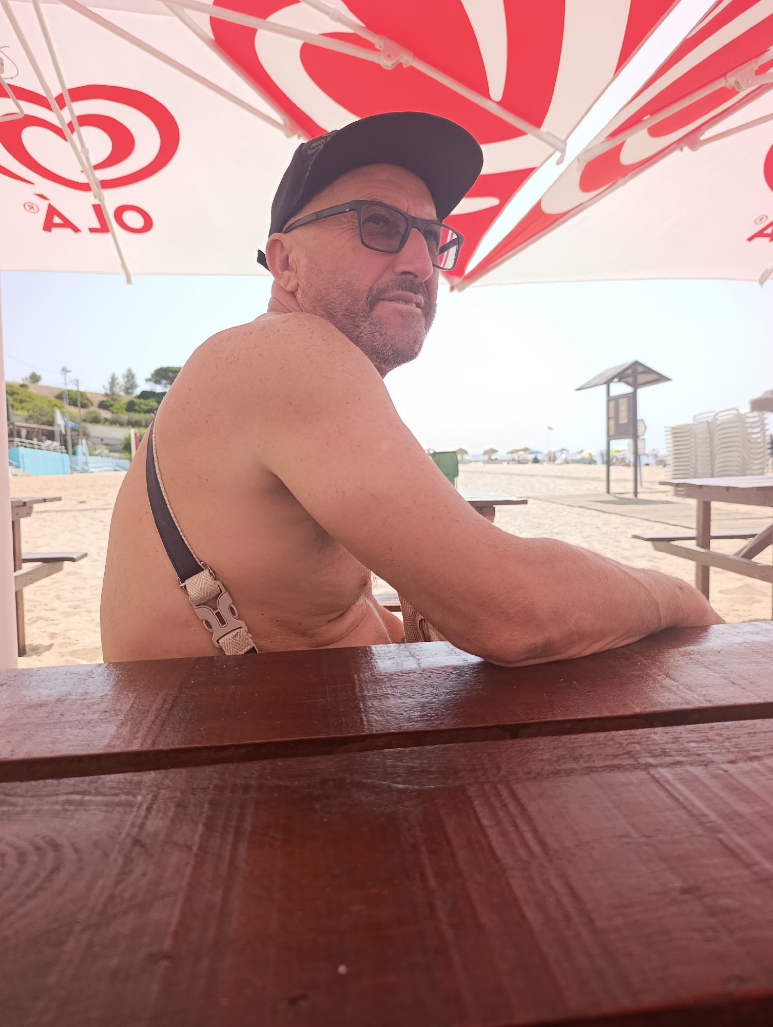 A man wearing a black baseball cap and glasses is sitting at a wooden table on a sandy beach under a large umbrella. The umbrella has a red and white striped pattern and a logo that reads "OLA". The man is shirtless, with dark hair and a beard, and appears to be in his 50s or 60s. His left arm is resting on the table, and a dark strap with a buckle is visible on his shoulder. In the background, there is a lifeguard tower, beach chairs, and umbrellas. The scene suggests a sunny day at the beach.