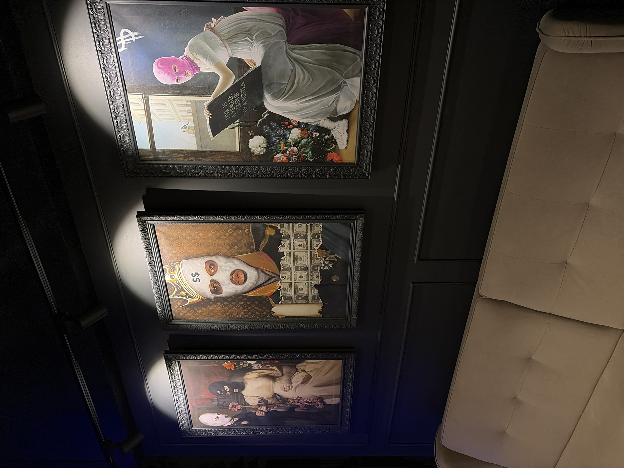 The image shows a wall with three framed art pieces hanging vertically. The top piece features a figure wearing a pink balaclava and a white dress, with a book that reads "Diva is a Female Version of a Hustla". The middle piece depicts a person in a balaclava with dollar signs, a crown, and money in the background. The bottom piece shows three figures with balaclavas holding flowers. The wall is dark, possibly black or dark grey, with raised panels. A light-colored couch is visible to the right. The location is in Chaikovsky, Russia.