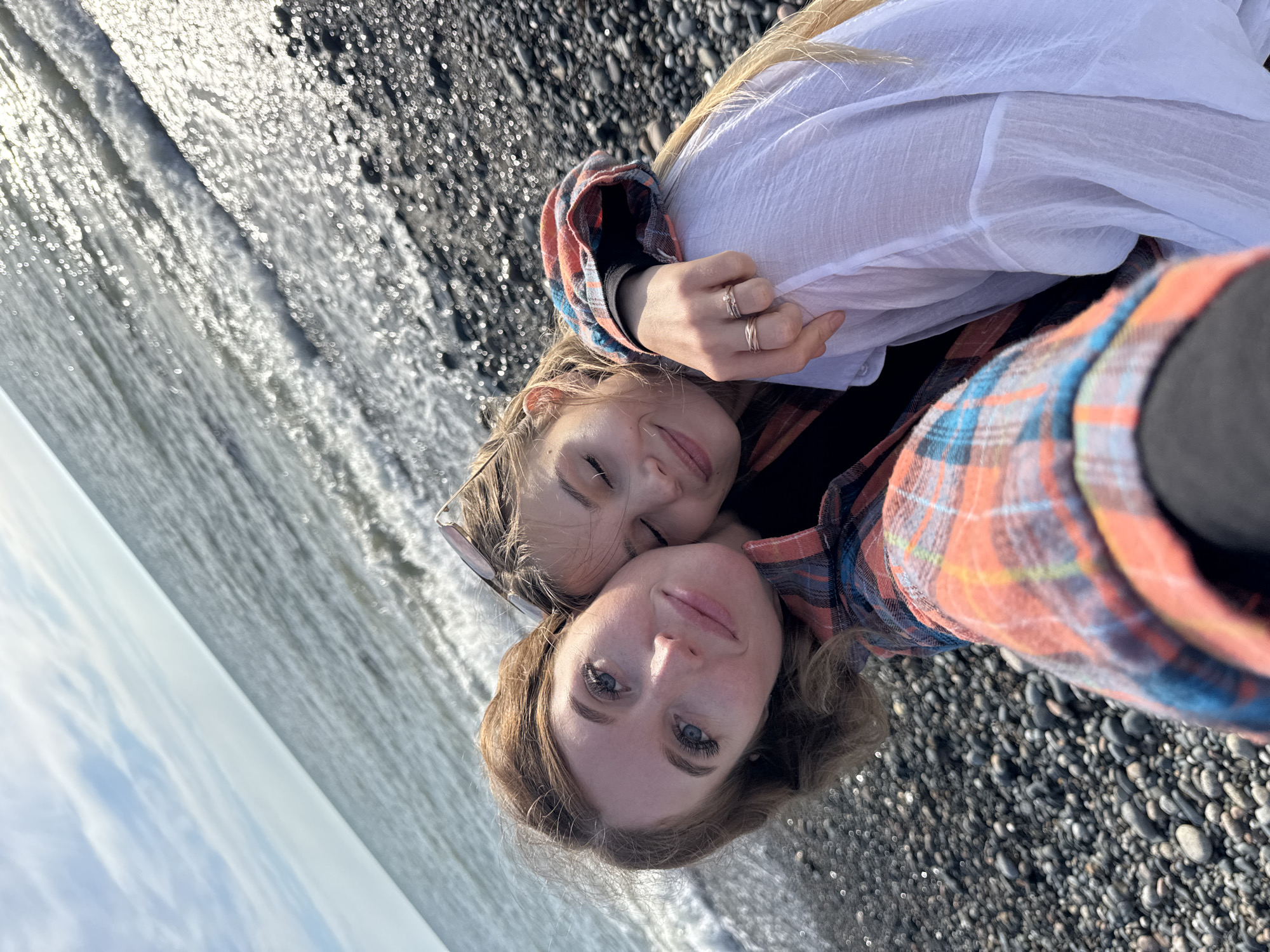 Two Caucasian women pose close together on a black pebble beach, likely taking a selfie. The woman in the foreground has light brown hair and blue eyes. The other woman, partially obscured behind her, has long blonde hair and is wearing sunglasses pushed up on her head. The background shows the ocean with shimmering sunlight reflecting off the water. The location is probably a beach near the city of Gulrypsh, Abkhazia, as indicated in the prompt.