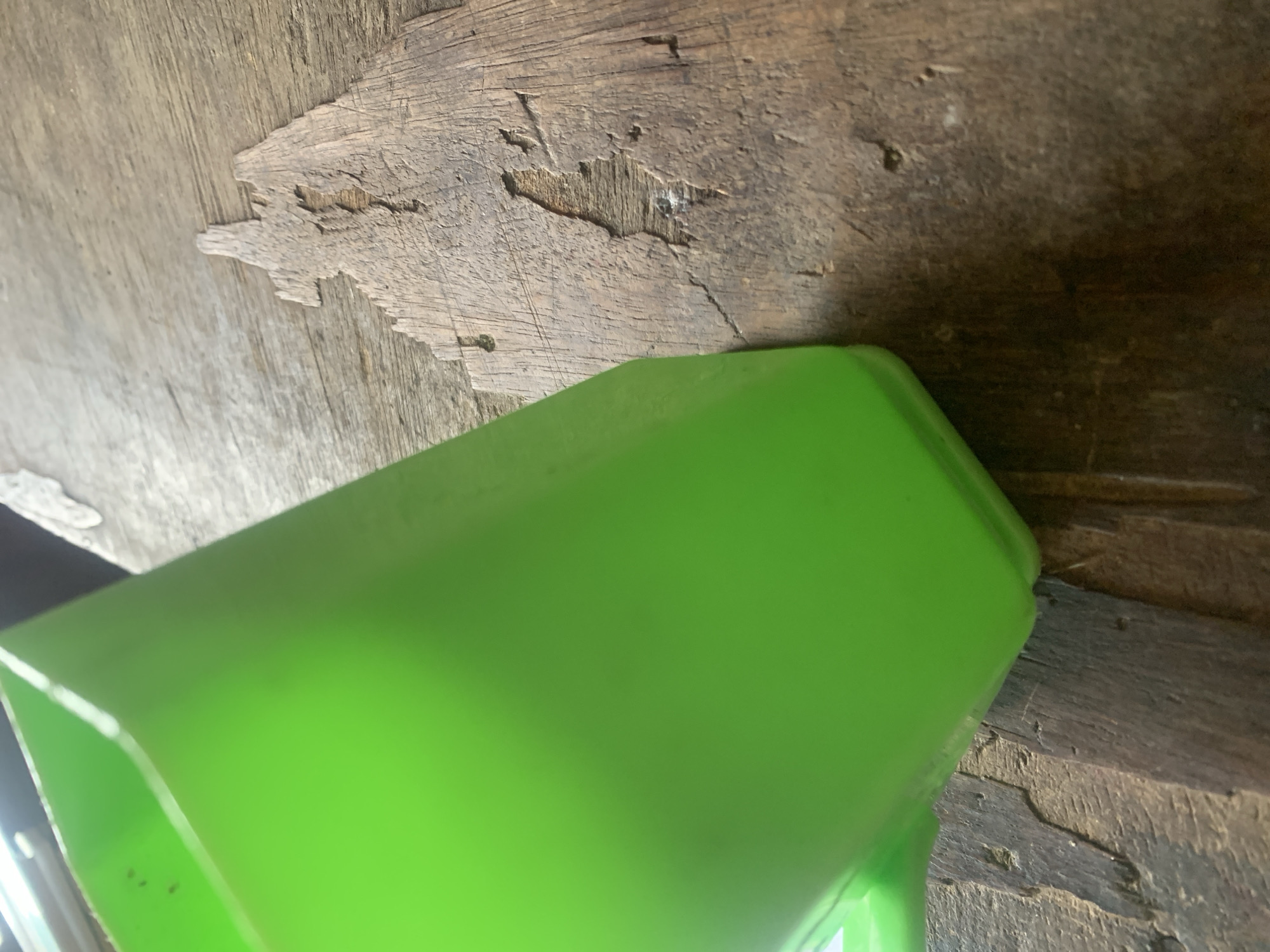 A bright green plastic cup is angled to the left of the frame, resting on a weathered wooden surface. The wood grain is visible, with areas of wear and tear that create an irregular texture. The background is mostly out of focus, with some lighter colored objects visible in the upper left corner. The image is taken from a close-up perspective, emphasizing the textures of the plastic cup and the wooden surface. The setting is an outdoor environment, likely a rough surface in Jalingo, Nigeria.
