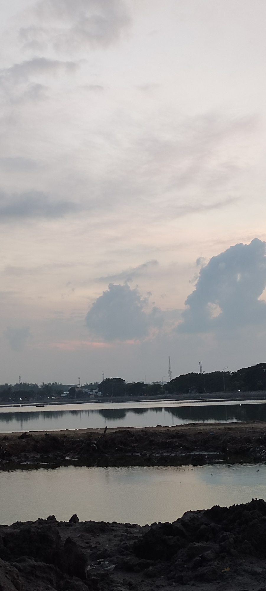 The image depicts a serene waterfront scene in Lhokseumawe, Indonesia. In the foreground, rough, dark soil and debris are piled high, creating a textured, uneven embankment. Beyond this, a calm body of water reflects the soft, muted colors of the sky.
In the middle ground, a narrow strip of muddy land separates two bodies of water. A single bird stands on this land, facing away from the camera. Further back, across a wider expanse of water, a dense line of trees and various structures form the distant shoreline. Among these structures are several tall communication towers, their slender forms reaching into the overcast sky. The water in the midground and background appears still, creating a mirror-like surface that reflects the faint outline of the trees and buildings.
The sky is predominantly gray with soft, diffused light, suggesting either early morning or late evening. There are scattered clouds, some appearing darker and more defined, while others are wispy and almost transparent, blending into the pale sky. A subtle pinkish hue is visible on the horizon, hinting at the twilight hours. The overall atmosphere is peaceful and quiet, with no clear signs of human activity other than the distant structures.