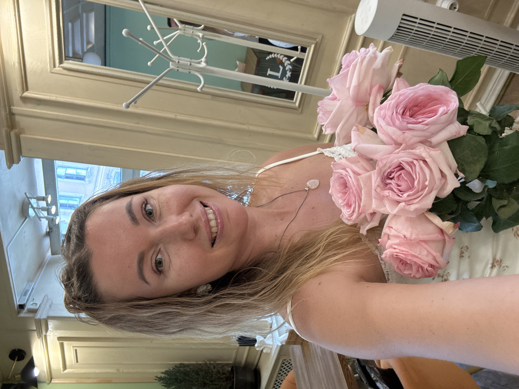 An indoor selfie captures an adult female holding a bouquet of pink roses. The woman, with light brown hair and fair skin, smiles directly at the camera. She is wearing a white tank top with lace detailing and a thin silver necklace with a heart-shaped, pale pendant. The bouquet consists of approximately five large, open pink roses with green leaves.
The background suggests an upscale cafe or restaurant setting. To the left, a dark green wall section is visible with a brass wall sconce, adjacent to cream-colored decorative wall paneling. A small potted evergreen plant rests on a table, partially obscuring a wooden table and an orange chair. Further left, a white decorative holder contains several dark, slender objects, and a white slatted radiator cover is present below.
To the right, more cream-colored wall paneling is visible, encompassing a large, ornate-framed mirror. The mirror's reflection shows a light green wall, a wooden table with a white 'L' and laurel wreath design, and a partial view of a person wearing a white shirt and dark trousers. A white, multi-hooked coat rack stands in front of the mirror. On the far right, a tall, white tower fan with a grilled surface is positioned vertically. The ceiling features light-colored panels and modern track lighting. The location is in the city of Unknown, Unknown.