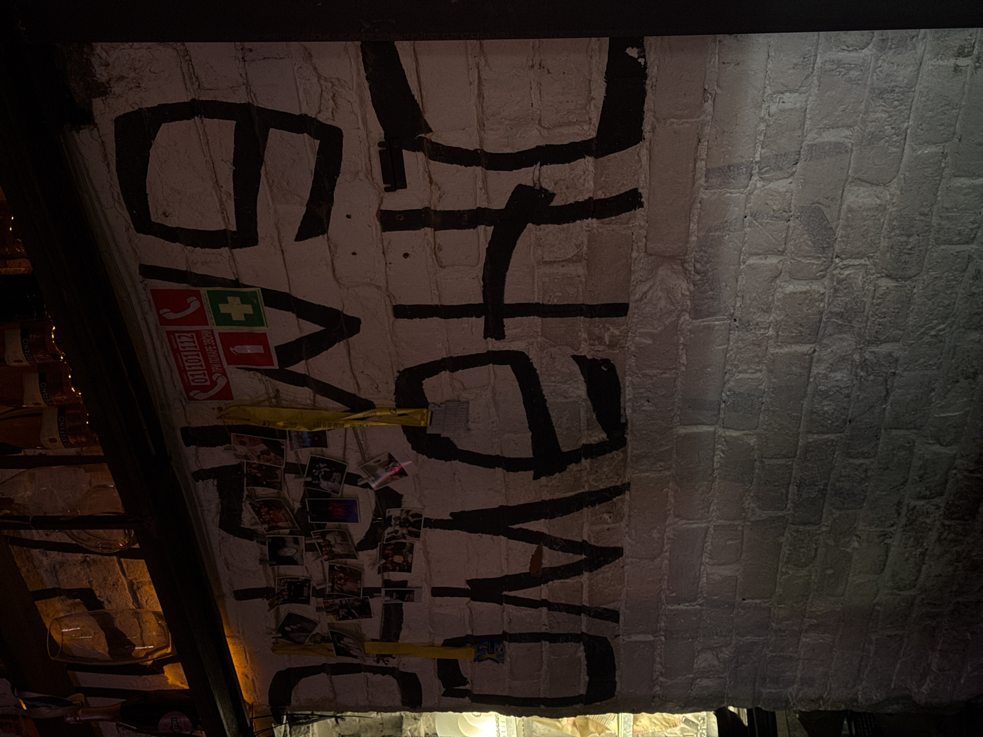 This image depicts a brick arched ceiling with white paint, and black painted lettering reading "HATE ME ON". Small photos are attached to the wall with yellow tape, along with safety signs. The location is Perm, Russia, based on the Cyrillic text on the safety signs. The focus is on the brick arch and lettering, with blurred edges of a room on the left-hand side, possibly a bar or cellar with some bottles and glasses.