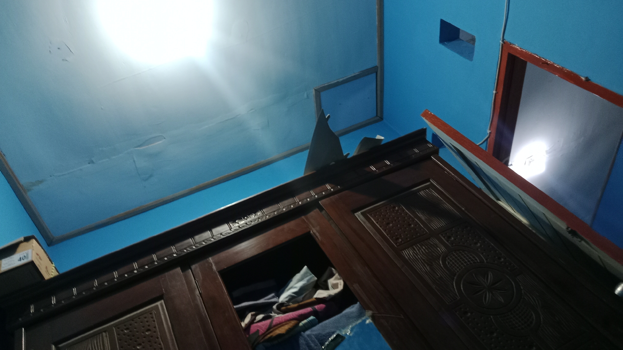 The image shows an interior view of a room with blue walls and a white ceiling. A dark brown wooden wardrobe with carved detailing is the central object in the foreground, with its doors slightly ajar, revealing stacked folded laundry. On top of the wardrobe, there is a cardboard box with the number "40" visible. In the background, there is a mirror with a reddish-brown frame, reflecting a bright light source, likely a ceiling light. A square opening or recessed light fixture is visible on the blue wall. The perspective is low, looking upwards. The scene appears to be inside a residential dwelling. This image is not specific enough to identify a location within Denpasar, Indonesia.