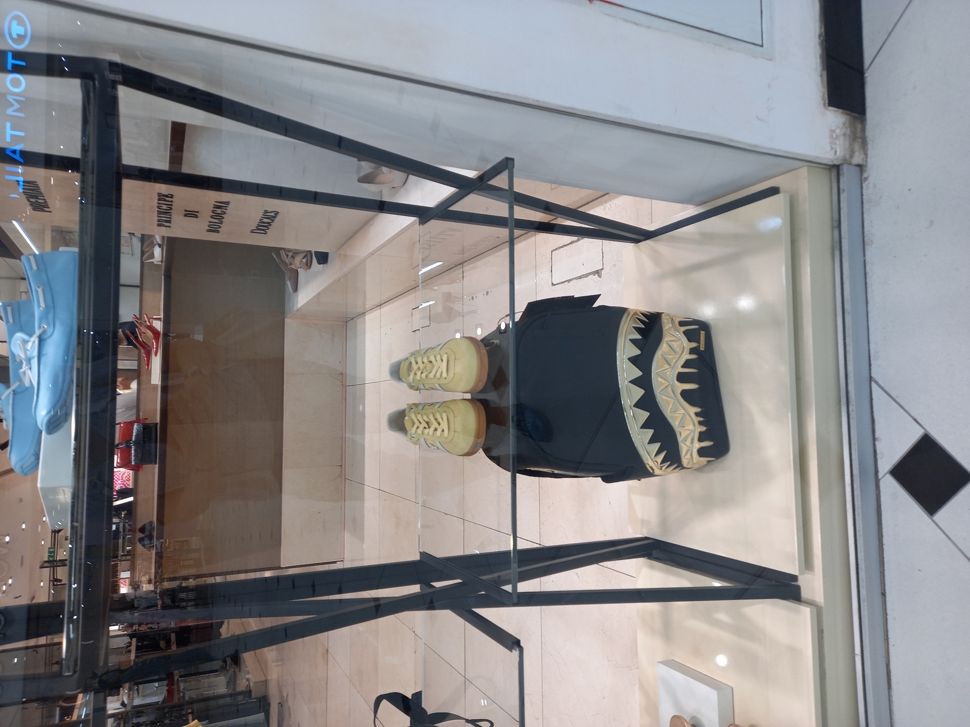 This is a medium shot of a store window display. The foreground is dominated by a black backpack with a gold shark tooth design. Placed on top of the backpack are a pair of yellow sneakers. The display is framed by a black metal shelving unit. In the background, more shoes and bags are visible on shelves and counters. A sign reads "PRNCIPE DI BOLOGNA DOUCAIS". The store appears to be located in Yekaterinburg, Russia, based on the text visible on the glass.