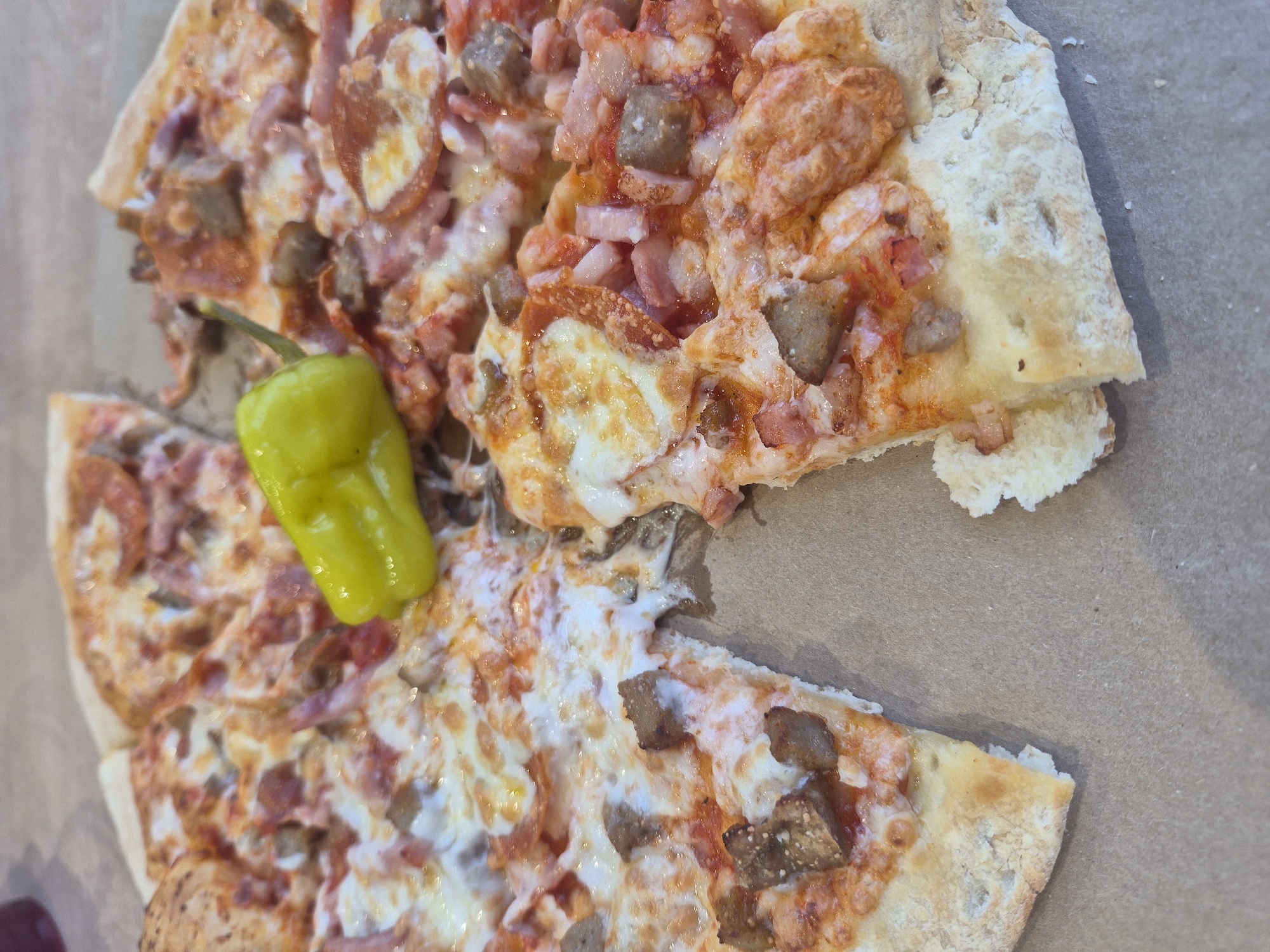 The image shows a partially consumed pizza with a yellow pepper on a brown surface. The pizza has toppings including sausage, ham, and cheese. The crust appears to be thick and uneven. The pepper is positioned near a cut portion of the pizza. There are no people visible in the image. The location is unknown.