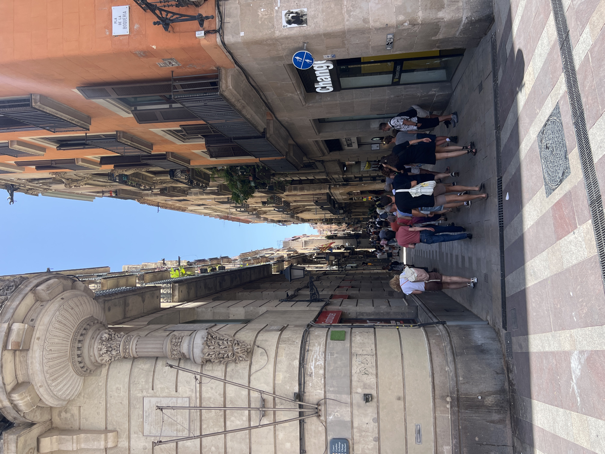 The image captures a narrow street in Barcelona, Spain, likely in the Gothic Quarter, given the architecture and the sign for "Pla de la Boqueria." The street is lined with tall, old buildings exhibiting a mix of architectural styles. The left side of the image features a large, ornate stone building with detailed carvings and a plaque. The right side of the street is dominated by an orange-colored building with balconies and shuttered windows. People are walking down the street, appearing to be tourists. Some are dressed in casual summer clothing. One person is wearing a white t-shirt and brown shorts, carrying a backpack. Another is wearing a red t-shirt and blue jeans. A sign for "Change" with Western Union branding is visible on the right side of the street, indicating a currency exchange service. The ground is paved with a distinctive pattern of red and white stripes. The sky is clear and blue, suggesting a sunny day.