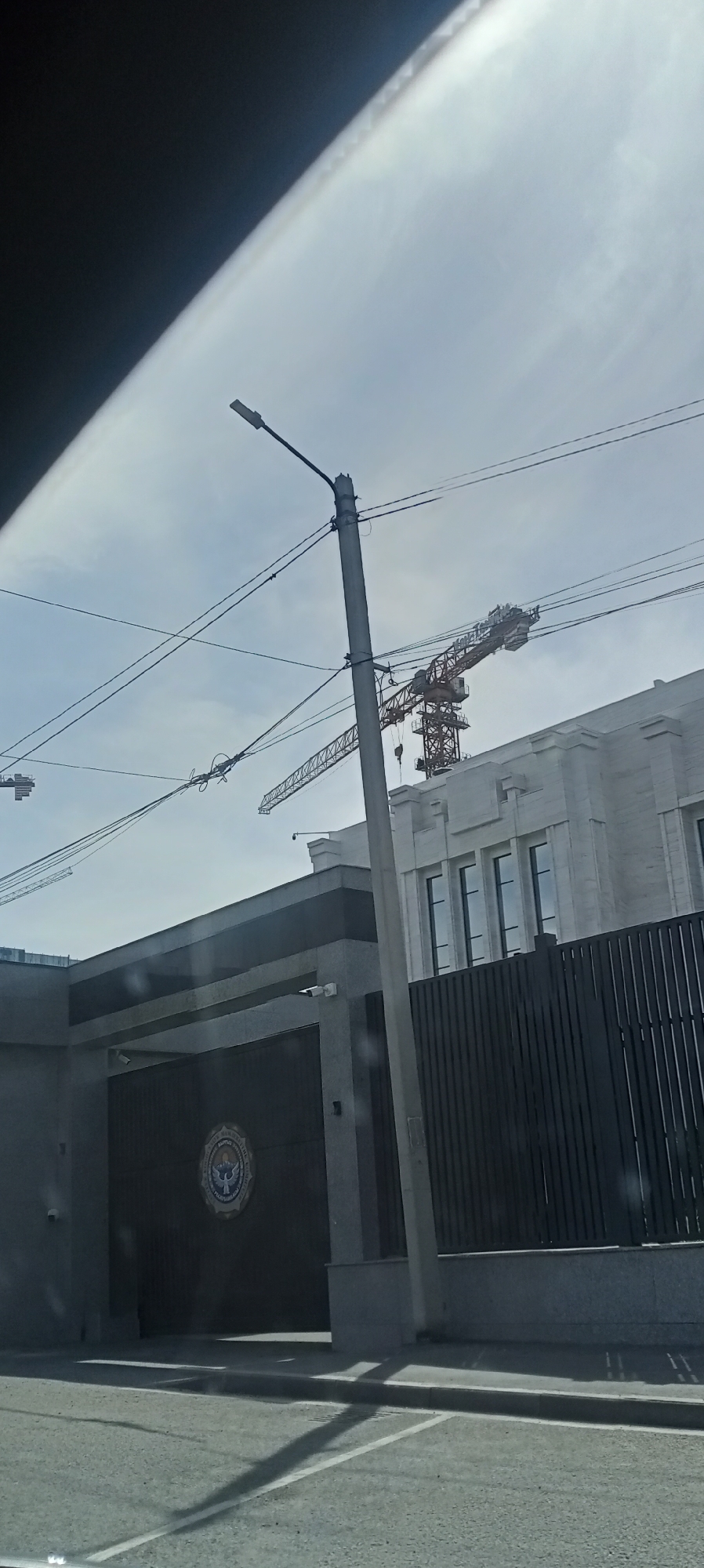 The image shows a view of the Ministry of Foreign Affairs of the Kyrgyz Republic building in Bishkek, Kyrgyzstan. The building is modern in design, with a white facade and large windows. A large, dark gate with a circular emblem is visible in the foreground. There are also two construction cranes in the background, suggesting ongoing development in the area. The sky is overcast, and the lighting is diffused. The image is taken from a street level perspective, likely from a moving vehicle, as indicated by the diagonal shadow on the road and the angle of the shot. A lamppost stands in front of the gate, and power lines are visible crisscrossing the sky.