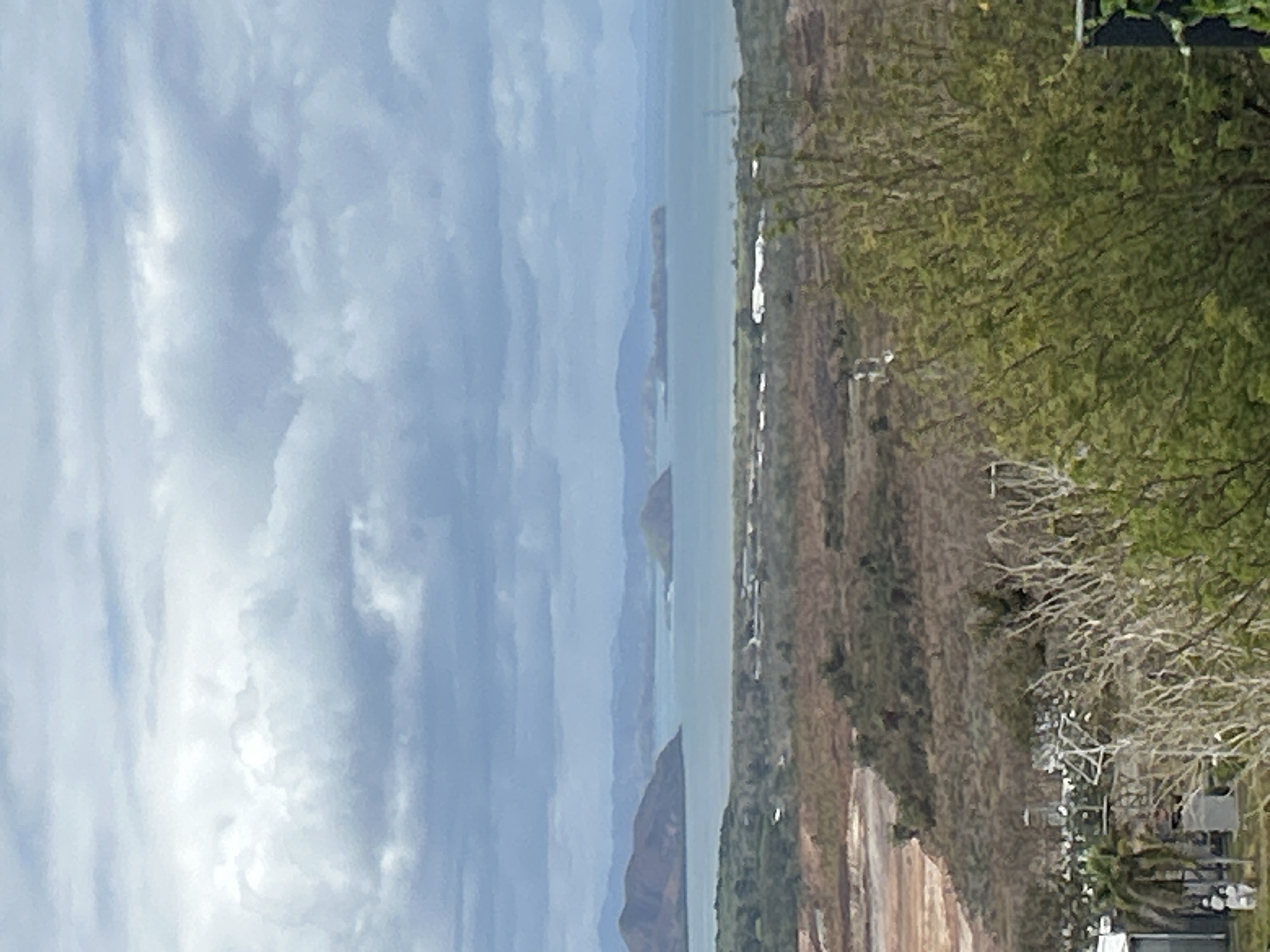 This image depicts a panoramic view of a coastal landscape, likely from an elevated vantage point within Port Moresby, Papua New Guinea. The foreground features dense, dry vegetation and some bare, branching trees. In the mid-ground, there is an area of disturbed earth with a reddish-brown hue, suggesting construction or excavation activity. Further back, a flat area with scattered buildings and structures, possibly a settlement or industrial zone, is visible along the coastline. The background is dominated by a body of water, likely the Fairfax Harbour or the Coral Sea, with several small islands and larger, mountainous islands in the distance. The sky is overcast with dramatic, layered clouds, indicating a potentially humid or changeable weather day. The overall impression is one of a developing coastal area with a blend of natural and man-made elements under a moody sky.