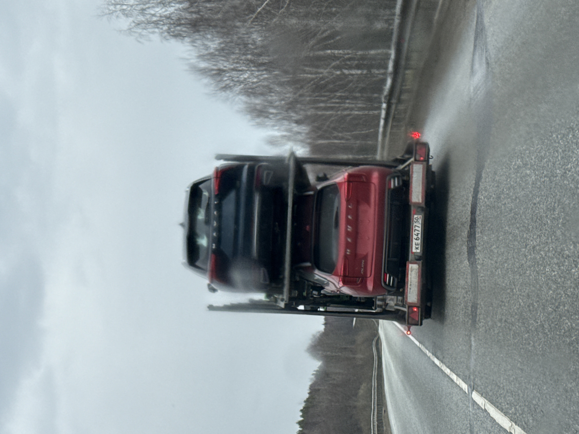 The image captures the rear view of a car carrier transporting two SUVs on a wet highway in Ust'-Syny, Russia, under an overcast sky. The carrier itself is partially visible, with its rear platform holding a dark-colored SUV or crossover on the upper level and a red SUV or crossover on the lower level. The red vehicle appears to be a Skoda Kodiaq, with faint, partially visible text that could be "KODIAQ" on its side.
The road surface is wet, suggesting recent rain or snow, and the surrounding environment features a treeline with bare trees on a gentle slope, indicative of a natural, possibly rural or semi-rural, setting. The sky is uniformly grey, suggesting a gloomy or wintery day.
A prominent detail is the license plate of the car carrier, displaying the Russian registration number "KE 6477 50". The "50" is the regional code for Moscow Oblast, Russia. The carrier also exhibits red taillights, which are illuminated. The scene depicts a typical commercial vehicle operation, transporting cars along a Russian road.