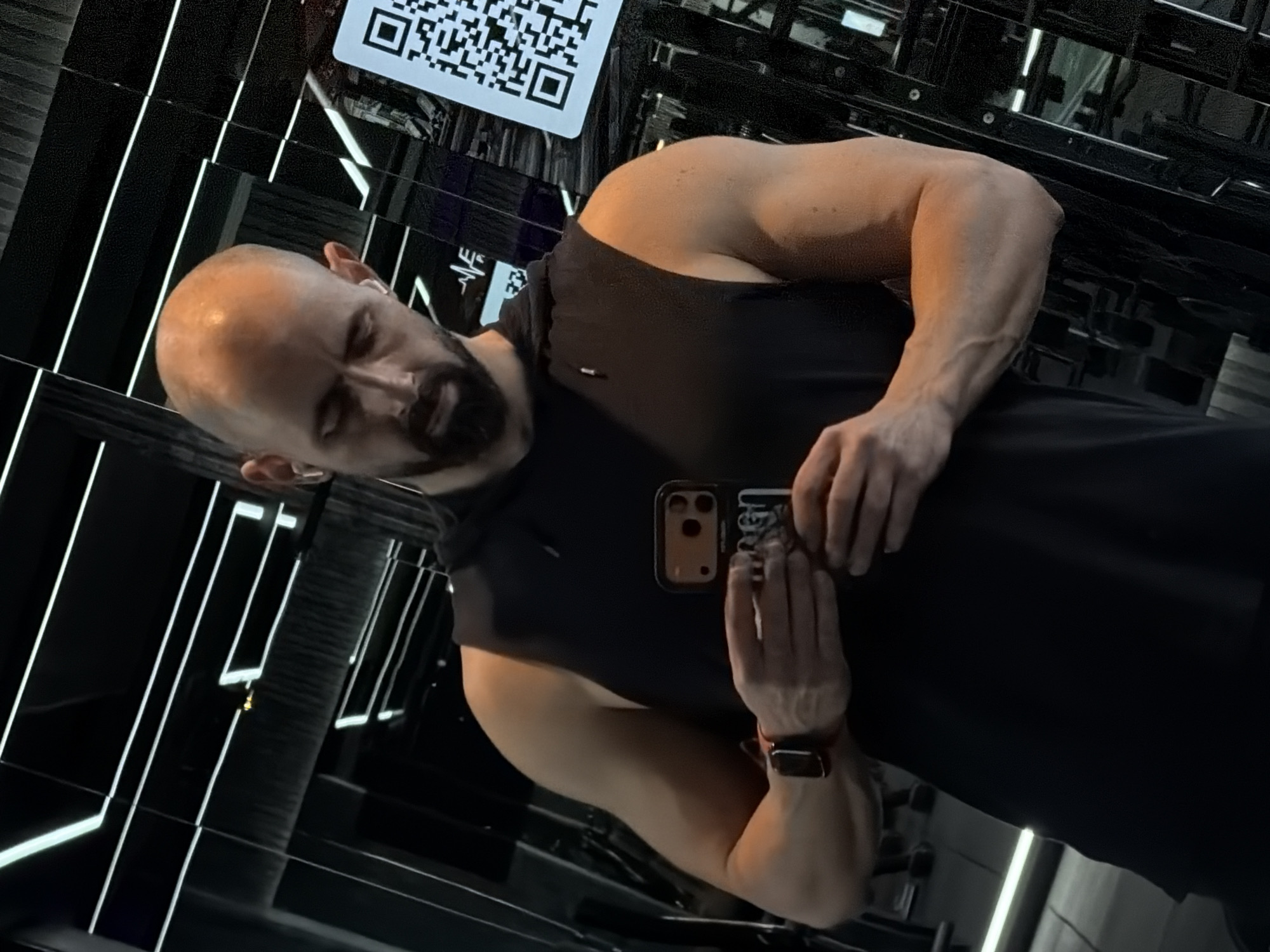 A bald man with a beard is taking a selfie in a gym in Plovdiv, Bulgaria. He is wearing a black tank top and a smartwatch. The gym has mirrored walls and a barcode displayed on one of the mirrors. Exercise equipment is visible in the background. The man is holding a phone with his left hand and posing with a slight smile.