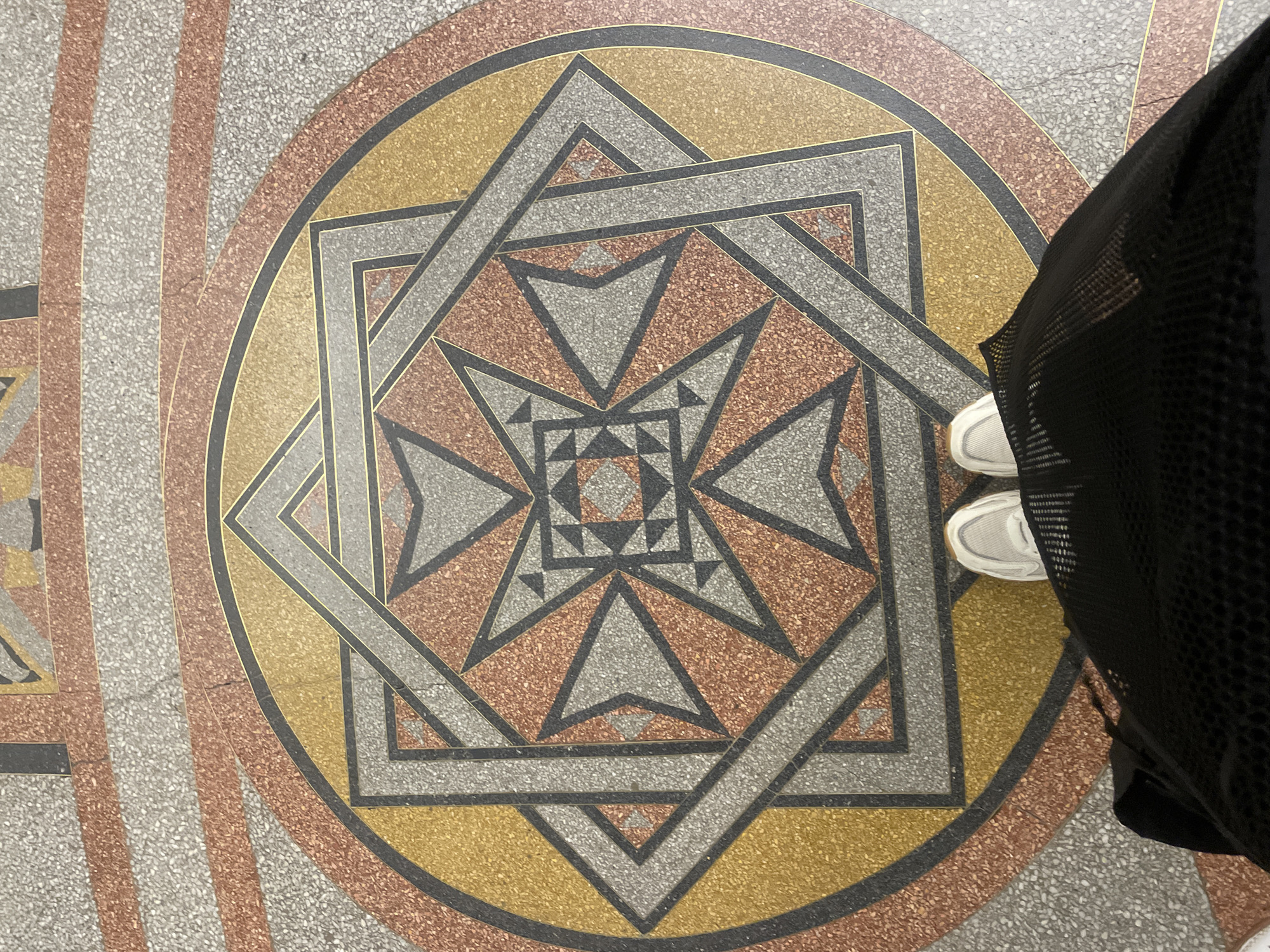 This is a top-down, medium shot image taken in Kronstadt, Russia, featuring a person standing on an ornate floor mosaic. The mosaic is circular and consists of concentric rings and geometric patterns. The outermost ring is a speckled pink and gray pattern, followed by a thinner gold ring, then a wider ring of alternating pink and gray speckled bands. Inside this, a large black circle encloses a complex geometric design. This design features interlocking squares and triangles in shades of gray, orange-brown, and gold, with thin black outlining. The center of the mosaic contains a smaller square with a triangular star pattern. The person is positioned in the lower part of the frame, standing with their feet on the mosaic. They are wearing white athletic shoes and a black, sheer, knee-length skirt. The perspective suggests the photo was taken by someone standing directly above or looking down at the person. The lighting appears even, likely from an overhead source.