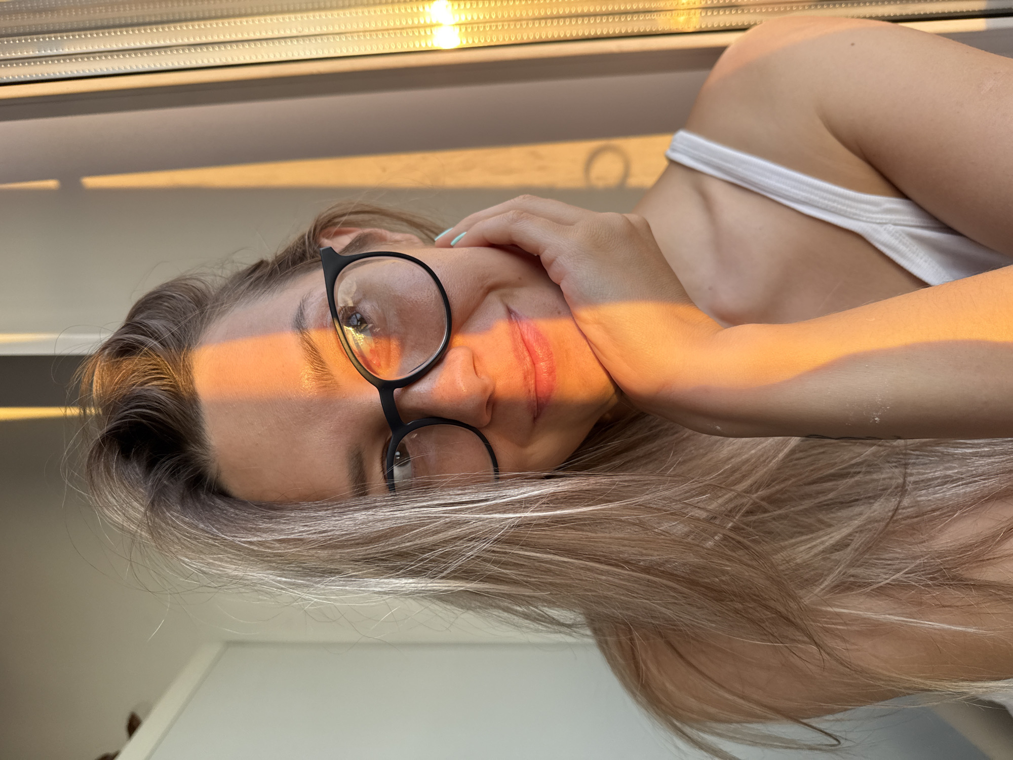 A young woman with blonde hair and glasses is looking to the side, with her hand resting on her chin. She is wearing a white tank top. The lighting suggests it is sunset, with warm orange light casting a stripe across her face. The background is blurred, with a window and possibly curtains visible. The setting appears to be indoors, by a window. There is no specific visual information in the image to determine a location in Chaikovsky, Russia.