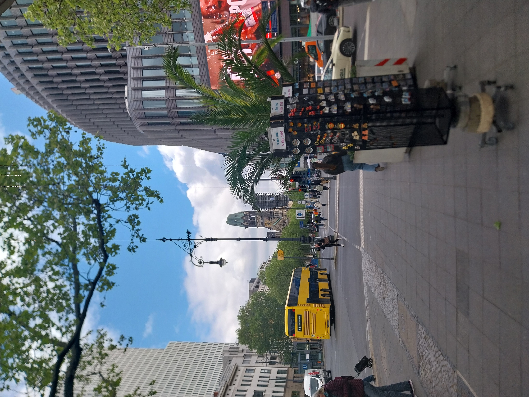 The image shows a street scene in Berlin, Germany. In the background, the Kaiser Wilhelm Memorial Church is visible. A yellow BVG double-decker bus travels on the street. A souvenir stand is on the sidewalk, offering items at prices of 4 Euros each, 3 for 10 Euros, or 5 for 15 Euros. Pedestrians are present. A person lies on the sidewalk in the lower right corner of the image. A modern building is on the left side of the street.