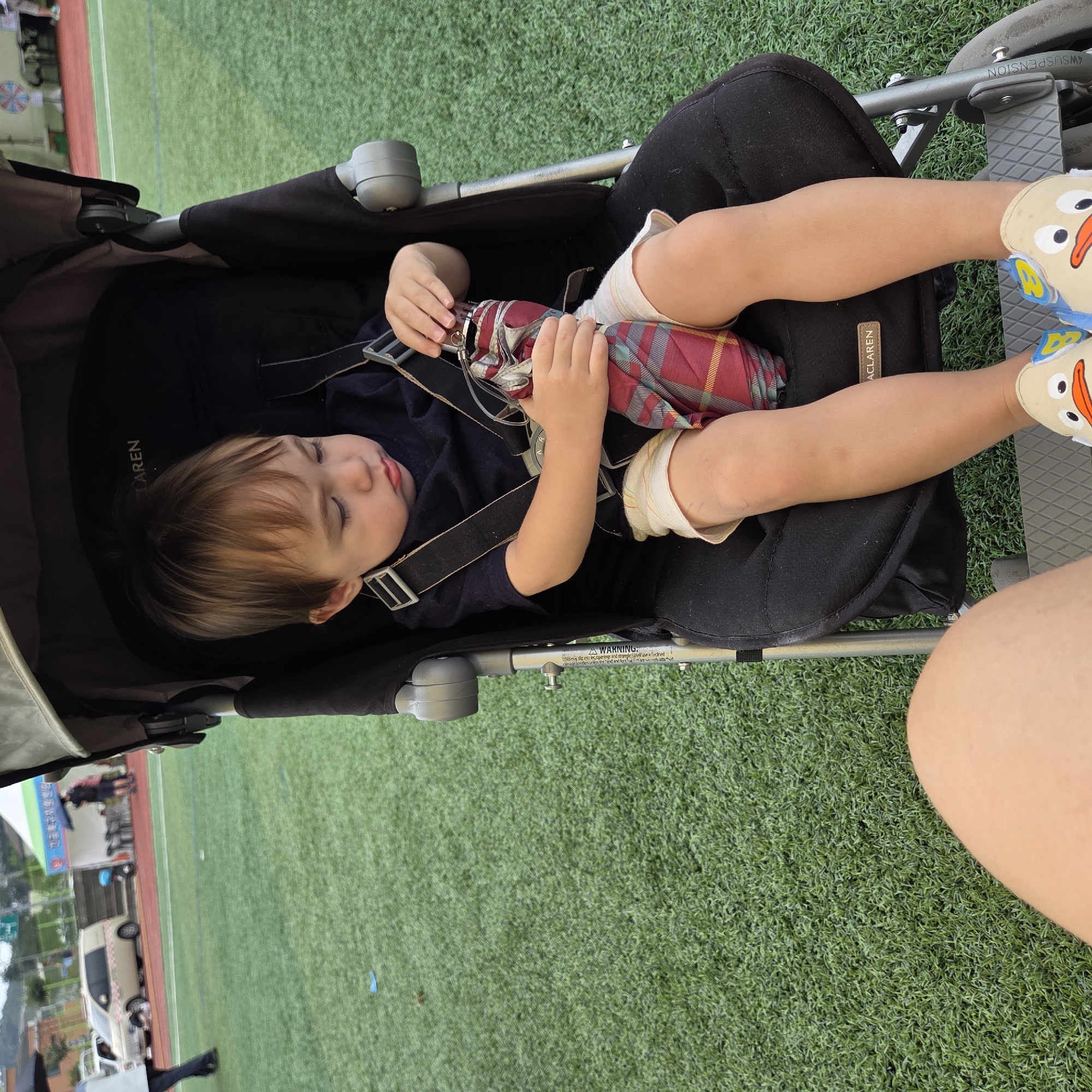 The image shows a toddler sitting in a stroller on a green turf field, likely a sports field, in Dunnae-myeon, South Korea. The toddler, with brown hair and wearing a dark blue shirt and light-colored shorts, is holding a folded umbrella with a red and green plaid pattern. The stroller is black and grey, with the brand name "MCLAREN" visible on the seat. The child's feet are visible at the bottom of the stroller, wearing light-colored slip-on shoes with a cartoon duck design. In the background, a tan-colored van is parked, and there are tents and some people in the distance. The overall scene suggests an outdoor event or gathering on a cloudy day.