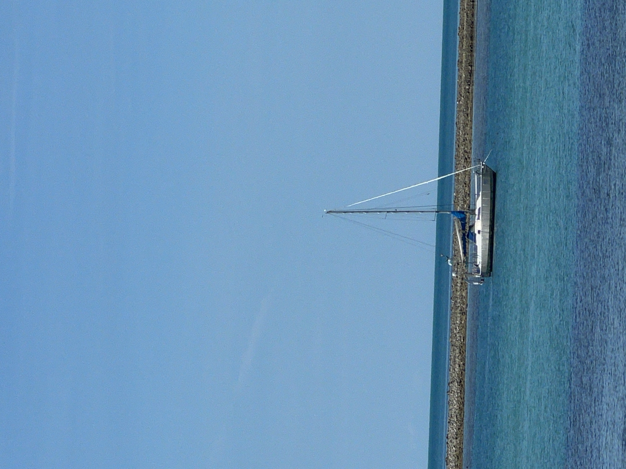 A sailboat is anchored in a calm, turquoise bay. The boat is white with a blue sail furled at the boom. A tall mast with rigging extends upwards, reaching into a clear, pale blue sky. In the background, a long, low breakwater made of grey stones runs horizontally across the scene. The water in the foreground is a deeper, darker blue, suggesting depth. The horizon line is sharp where the water meets the sky. The image appears to be taken from a slightly elevated vantage point, looking out over the water. The overall impression is one of peace and tranquility. This scene is located in Baan Hua Thanon, Thailand.