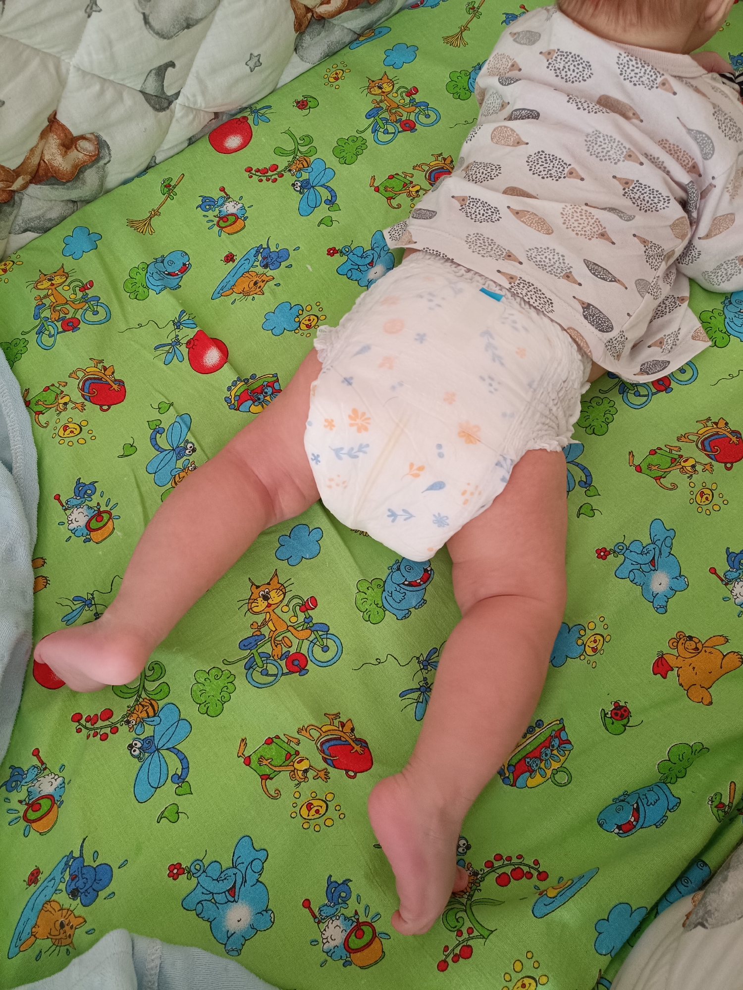 A baby lies prone on a green, patterned sheet in a crib. The baby wears a white shirt with hedgehog designs and a disposable diaper patterned with flowers and blue leaves. The sheet features colorful cartoon drawings of animals like cats on bicycles, hippos, elephants, bears, and dragonflies. The location is not specified within the image and cannot be determined to be Tobolsk, Russia.