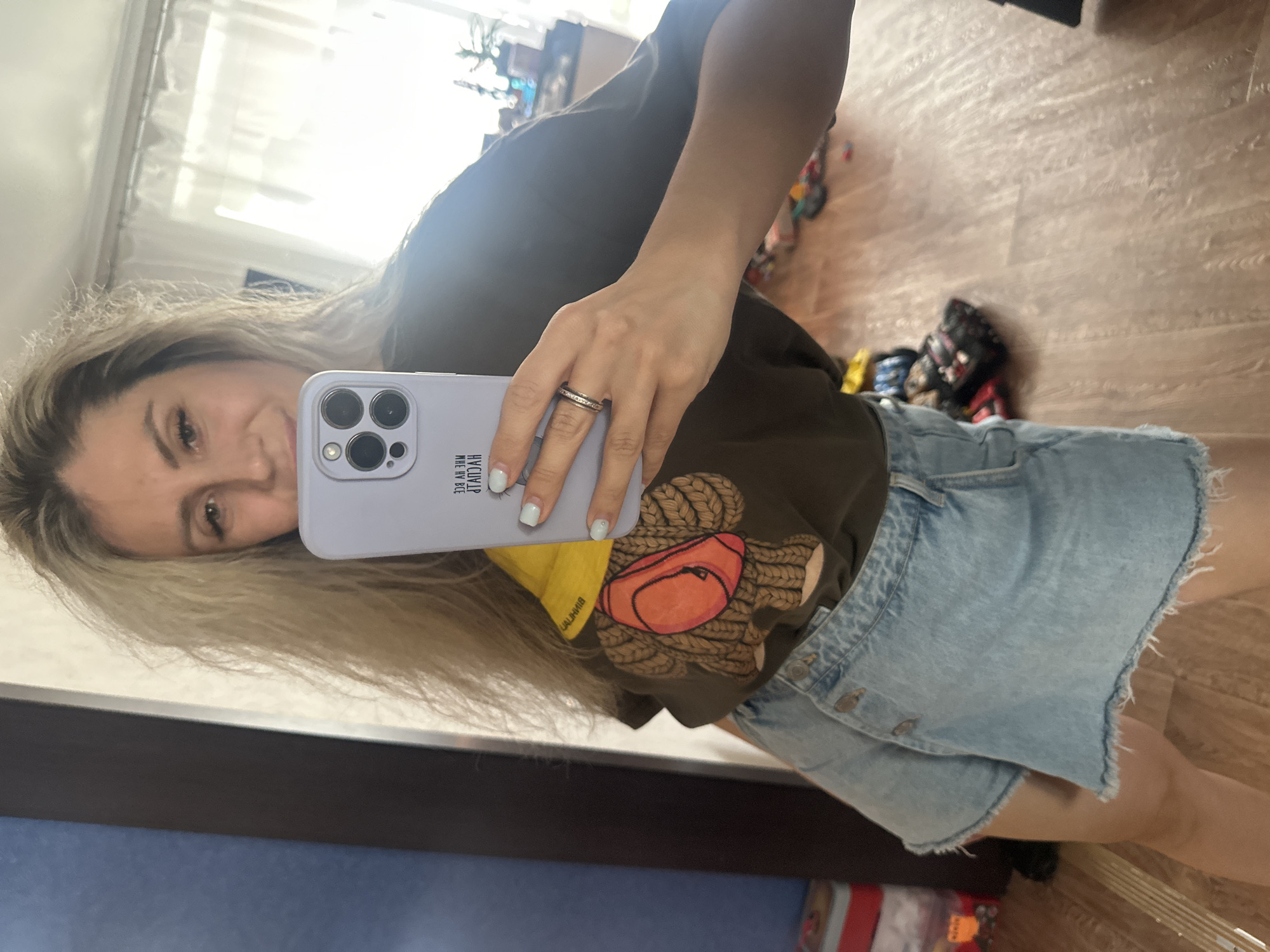 A young woman with long, messy blonde hair takes a mirror selfie indoors. She is wearing a dark green t-shirt with a cartoon bear graphic and a light blue denim skirt. She is holding a white phone in her right hand, which displays text reading "MHE HV BCE GTAH". She has on a ring with small stones on her left hand. The background appears to be a room with a wooden floor and a large mirror reflecting the scene. Other objects, possibly shoes and storage containers, are visible on the floor and in the background. The location is identified as Novoaltaysk, Russia.