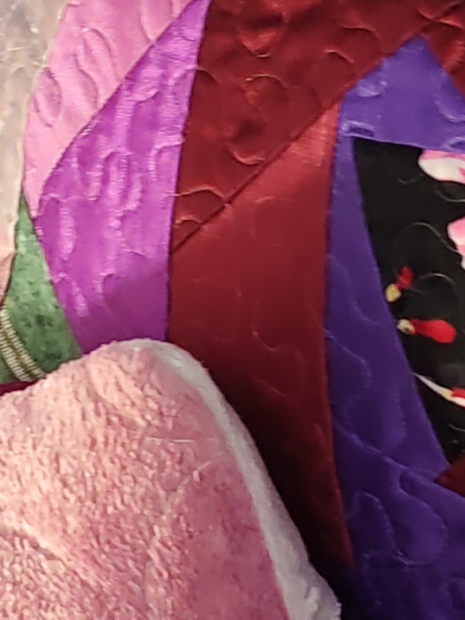 The image displays a close-up, abstract view of fabric pieces stitched together. The dominant colors are various shades of pink, purple, and deep red, with a patch of black fabric featuring a floral pattern in pink and red visible on the right side. Some of the fabrics have a quilted texture with wavy patterns. In the foreground on the left, a fluffy, light pink material is partially visible. The scene appears to be a detail of a textile craft or decoration. There is no discernible urban environment or location in Olginskaya, Russia, depicted in this image.