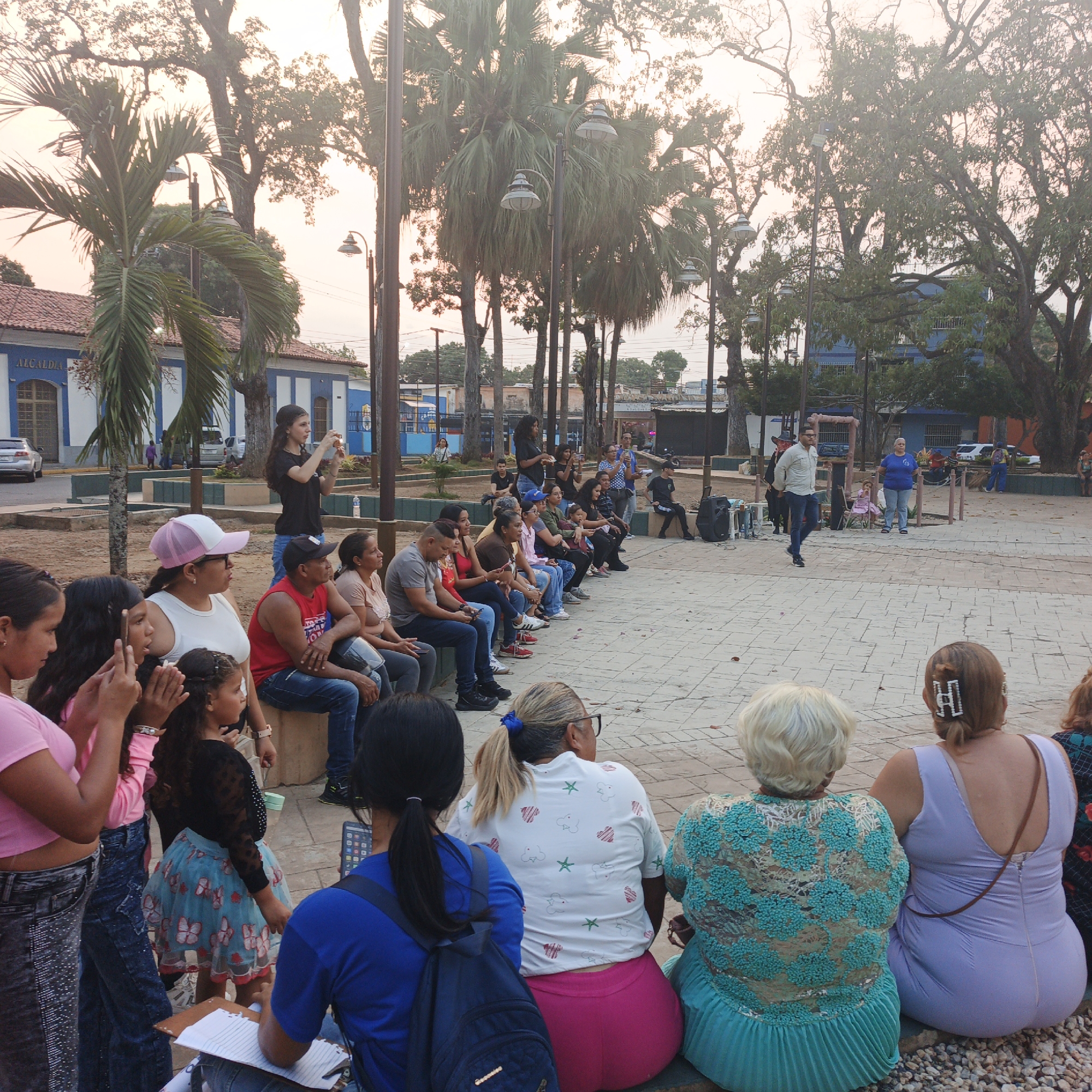 The image depicts an outdoor public gathering in a plaza or park situated adjacent to the Alcaldía (Mayor's Office) building in Tinaquillo, Venezuela. The Alcaldía building, a two-story structure with blue and white walls and a red-tiled roof, is visible in the background on the left. The scene is illuminated by soft, warm light, indicative of late afternoon or early evening.
Approximately 30-40 individuals of varying ages, predominantly adults, are present. In the immediate foreground, with their backs to the camera, are four women: one with light blonde hair wearing a teal top with intricate embellishments and a pleated skirt; one with reddish-brown hair in a light purple, v-neck dress featuring a horizontal hair clip; one with blonde hair in a ponytail, wearing glasses, a white t-shirt with scattered patterns, and pink pants; and one with dark hair, a blue t-shirt, and a dark backpack, holding a notebook and pen.
To the left, a group of approximately 15-20 people, including adults and several children, are seated in a line on a low concrete barrier, observing an activity in the central paved area. Notable individuals in this group include young girls in pink tops, some clapping or holding smartphones; a girl in a black long-sleeved top and a blue skirt with butterfly patterns; a woman in a white tank top and a pink and white baseball cap; and a man in a red tank top and blue jeans.
In the central paved area, a man in a light-colored long-sleeved shirt and blue jeans walks towards the left. A woman in a blue t-shirt stands near a wooden fence structure, while a small child in a pink dress sits on a chair behind it. A large black public address speaker is positioned on the right edge of the paved area. Further back, a woman in a black t-shirt is actively recording or photographing the event with a smartphone.
The environment includes numerous large trees, some palm trees, and several streetlights. A silver sedan is parked on the street behind the seated crowd. The ground surface consists of light-colored interlocking pavers.