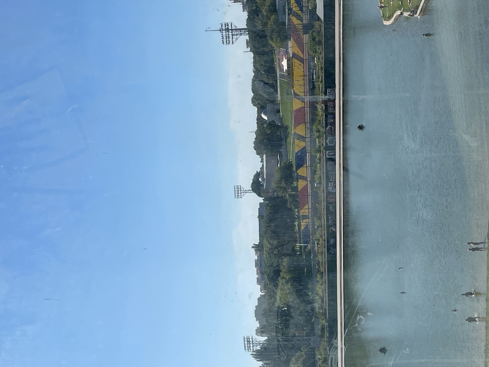 This is a medium-distance shot of a wide, calm body of water, likely a man-made reservoir or lake, at the Luzhniki Olympic Complex in Moscow, Russia. The water's surface reflects the clear blue sky and the surrounding greenery. In the foreground, the shallow edge of the water is visible, with several people wading and standing. Further out in the water, a few more individuals are swimming or floating.
In the middle ground, a long, raised embankment runs along the far side of the water. This embankment is adorned with vibrant graffiti and colorful, stadium-like seating, with sections of yellow, red, and blue. Behind the embankment, dense green trees and foliage create a natural barrier, partially obscuring the urban landscape beyond. Several tall, industrial-looking towers with numerous lights are positioned within the tree line and on the horizon, characteristic of sports stadiums or infrastructure lighting. The sky is a bright, cloudless blue, indicating a sunny day. The image appears to have been taken through a window, as evidenced by faint smudges and reflections on the surface.