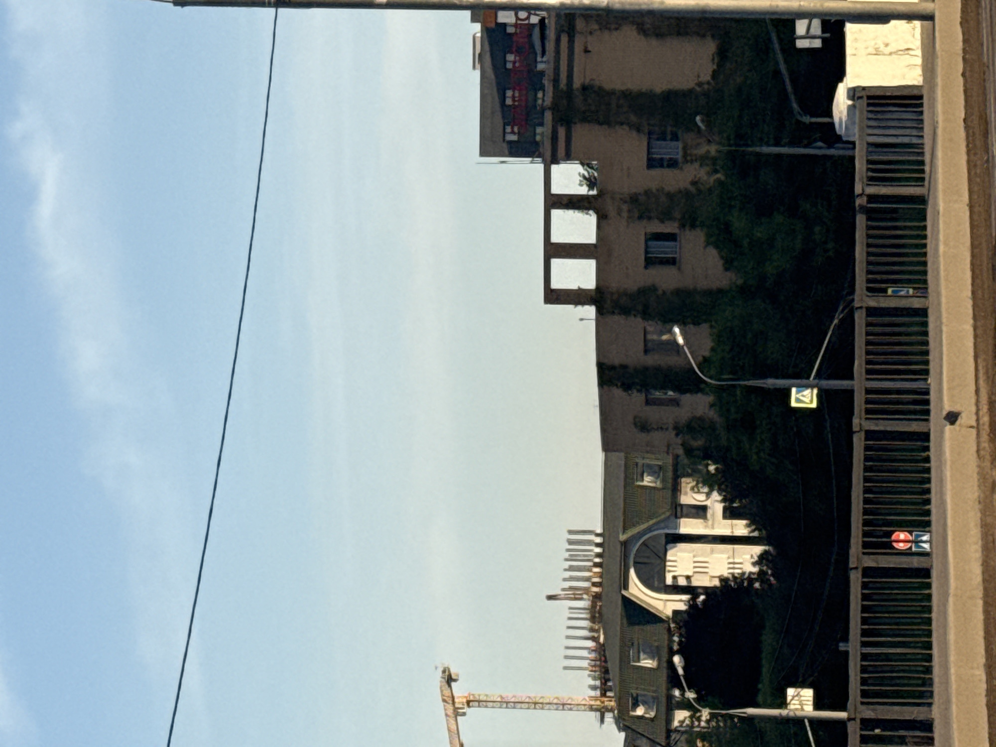 The image depicts a street scene in Khimki, Russia. The sky is visible on the left, transitioning to the facade of buildings on the right. The buildings appear to be older residential structures, some covered with vegetation. A construction crane is visible near one of the buildings, indicating ongoing construction or renovation work in the area. A streetlight, power lines, and a pedestrian crossing sign are also visible.