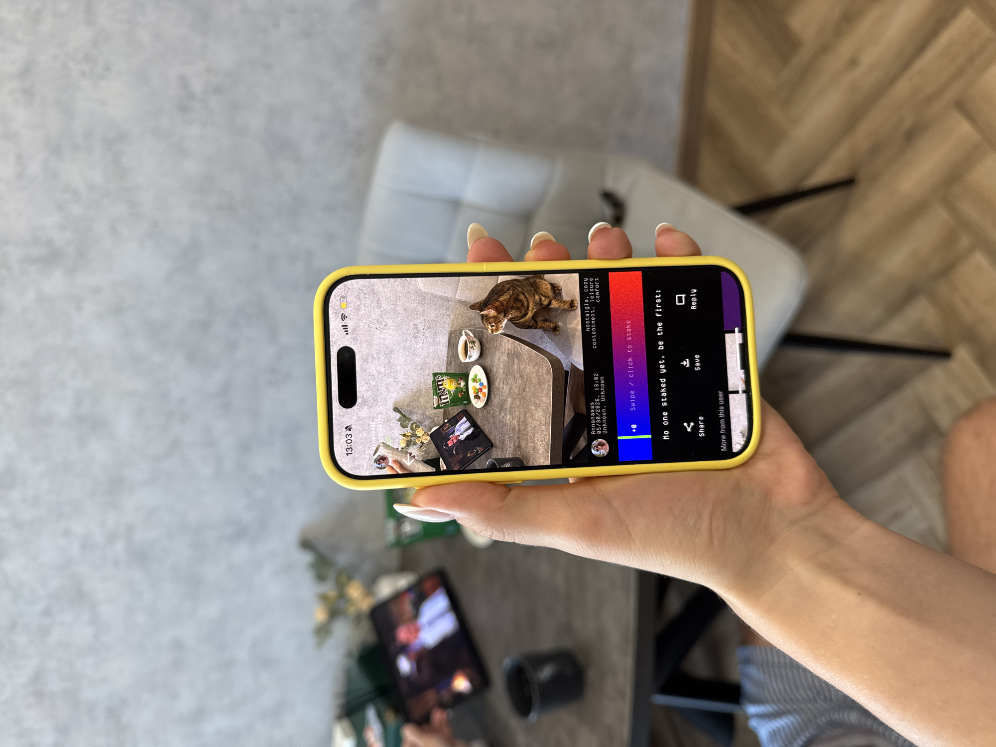 The image shows a hand holding a yellow-cased iPhone, displaying a social media post. The post, seemingly by "Annanaaas" on 05/30/2026 at 13:02 in Unknown, Unknown, features a cat on a table alongside items like M&Ms and a tablet. Text within the post includes keywords like "nostalgia, cozy, contentment, leisure, comfort" and calls to action like "Swipe / click to stake" and "No one staked yet, be the first". The options to share, save, and reply are also visible. An additional tablet displaying an image is seen in the blurred background, along with a cup and plants on a table. A chair is in the background as well.