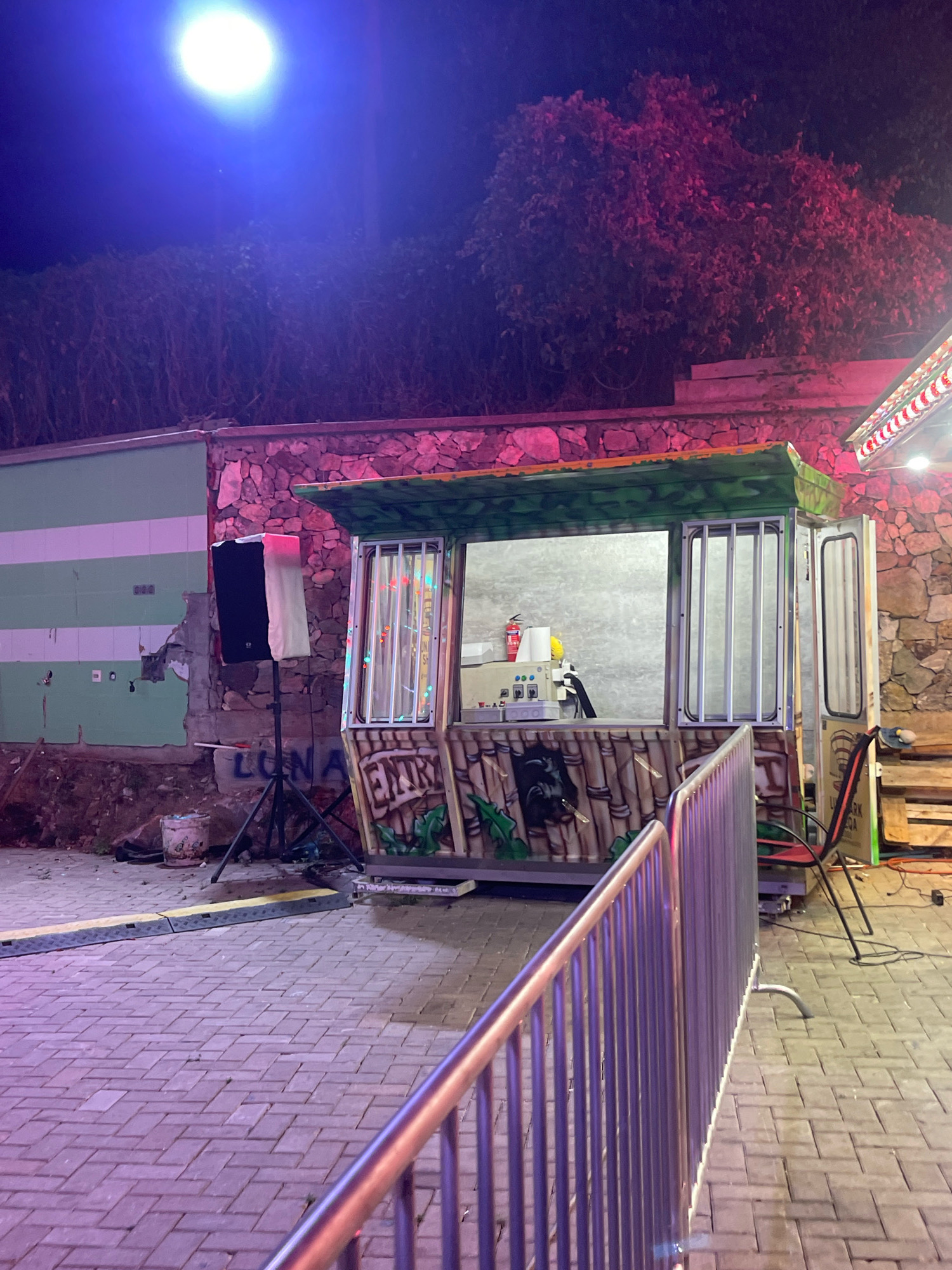 The image shows a nighttime scene in Prizren, Kosovo, featuring a small, decorated booth or stand. The booth has a green, leafy-patterned roof and a facade designed to look like bamboo, with an image of a black panther. It has two open windows for serving. In the foreground, a metal railing separates the viewer from the booth. Behind the booth, a stone wall is visible, illuminated by pink and purple lights. To the left of the booth, a speaker on a stand is positioned against a wall with green and white stripes. The ground is paved with interlocking bricks. A bright, circular light source, possibly a streetlamp, is visible in the upper left corner of the image, casting a blueish-purple glow. The overall impression is of an amusement park or fairground attraction.