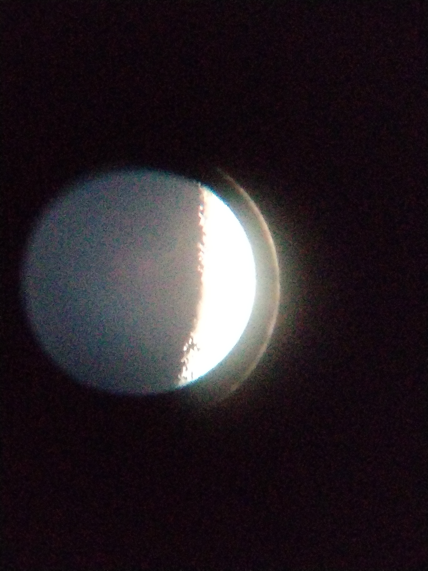 The image shows a close-up of a celestial body, likely the moon, in its crescent phase. The left side of the celestial body is dimly lit and appears mottled, possibly due to atmospheric interference or the resolution of the imaging device. A bright, sharp line of light delineates the terminator, the boundary between the illuminated and unilluminated portions of the celestial body. The right side of this illuminated portion is intensely bright, almost blown out, suggesting a direct view of the sunlit surface. The background is entirely dark, consistent with an astronomical observation. There are no identifiable landmarks or environmental features to indicate a specific location in a city or otherwise.
