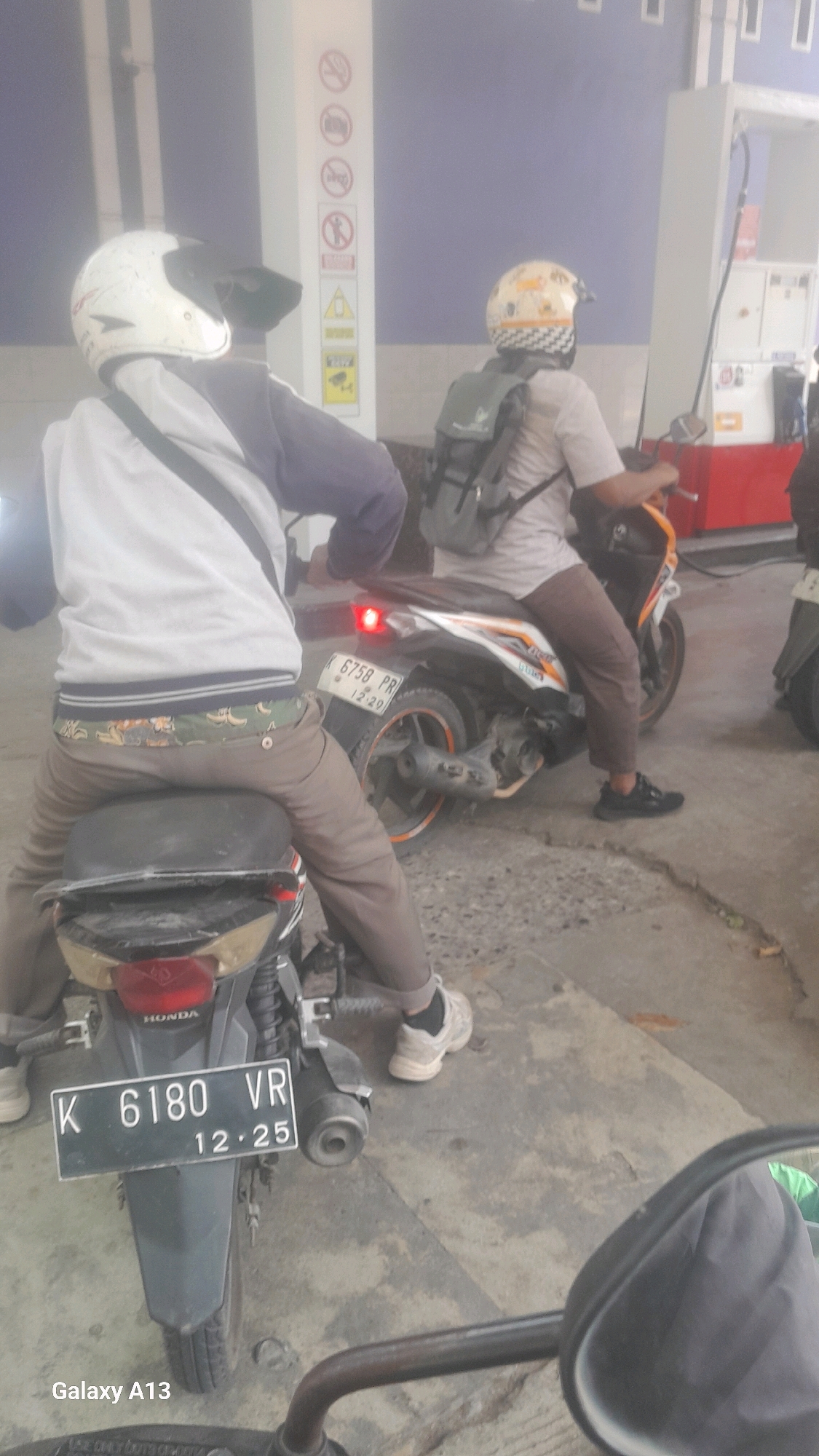 Two individuals on motorcycles are at a gas station. The person in the foreground, wearing a white helmet and a light-colored jacket over a darker shirt, sits on a motorcycle with a license plate reading "K 6180 VR 12.25." This motorcycle's rear is prominently featured. The second individual, wearing a patterned helmet and a light-colored shirt, is on a motorcycle in the background, facing towards a gas pump. This motorcycle has a backpack attached to its rear. The gas station has blue walls and visible safety signage, including a "no smoking" symbol. The ground is paved concrete. The image is taken from a low angle, possibly from another motorcycle. The presence of Indonesian license plates suggests the location is in Indonesia, specifically Tanjunggemuk, Indonesia, as indicated by the license plate prefix "K" which corresponds to the Keduang region of Central Java, Indonesia.