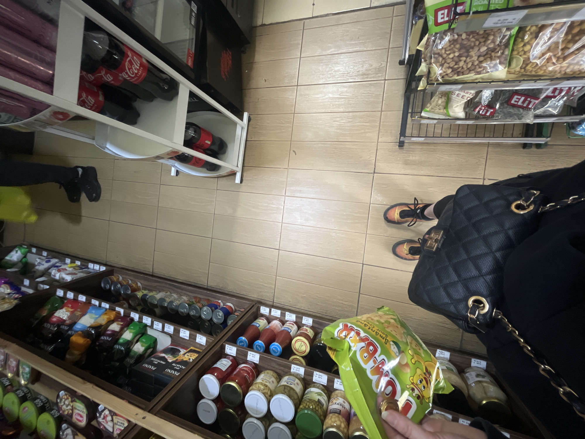 The image shows a view inside a shop, presumably in Sofia, Bulgaria, given the presence of Bulgarian language product labels on the shelves. The floor is tiled with light brown tiles.
A person is holding a bag of chips, labeled "Smântână Sour Cream" in Bulgarian. They are standing near shelves displaying various products, including canned goods, sauces, and bottled drinks.
To the left, a shelving unit holds various bottles of Coca-Cola. A black shopping bag and a portion of the person's clothing are visible, along with their feet and laced shoes. A separate rack holds various types of nuts, including ELITE brand products.