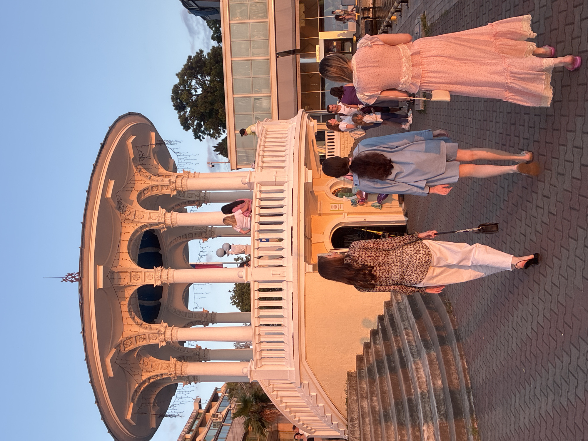 The image depicts an outdoor scene at sunset in Sochi, Russia, likely on the waterfront. In the foreground, three women are walking away from the camera on a brick-paved pathway. The woman on the left wears a patterned long-sleeved shirt and white pants. The middle woman wears a blue oversized shirt and shorts. The woman on the right wears a pink floral dress.
Behind them, a prominent white rotunda with classical architectural elements, including columns and arches, is illuminated by the warm light of the setting sun. The rotunda has a balustrade around its upper level, and people can be seen standing on and around it. In the background, buildings, trees, and a clear blue sky are visible. The overall atmosphere suggests a pleasant evening on a promenade.