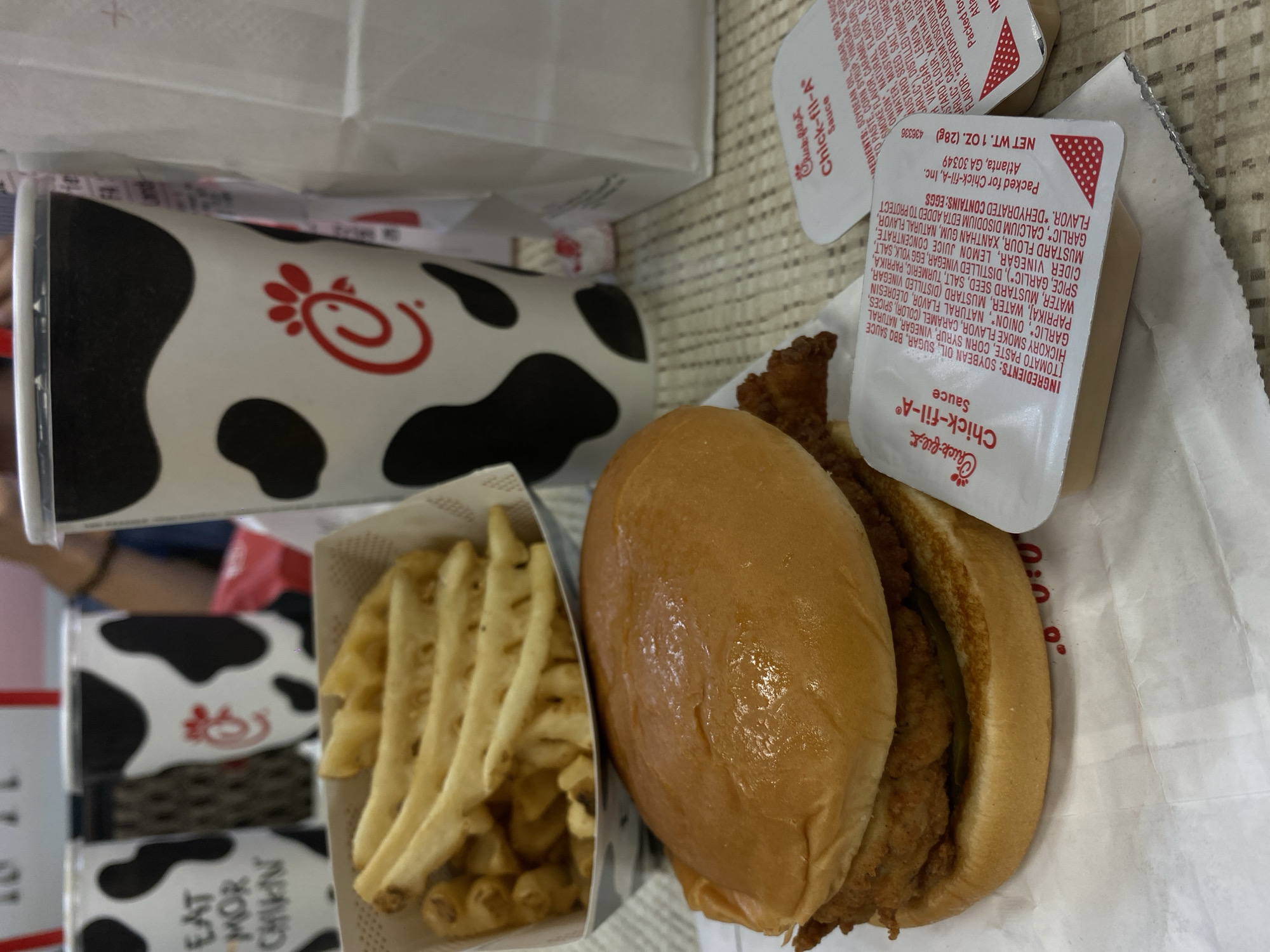 This is a close-up, eye-level, medium shot of a Chick-fil-A meal on a table in Hagerstown, Maryland. In the foreground is a chicken sandwich on a bun, with waffle fries in a white paper container behind it. Two Chick-fil-A sauce packets are next to the sandwich. In the background, there are several large Chick-fil-A branded cups with a cow print design. One of the cups has a red straw. The table appears to be a light-colored wood. The overall scene suggests a casual dining experience at a fast-food restaurant.