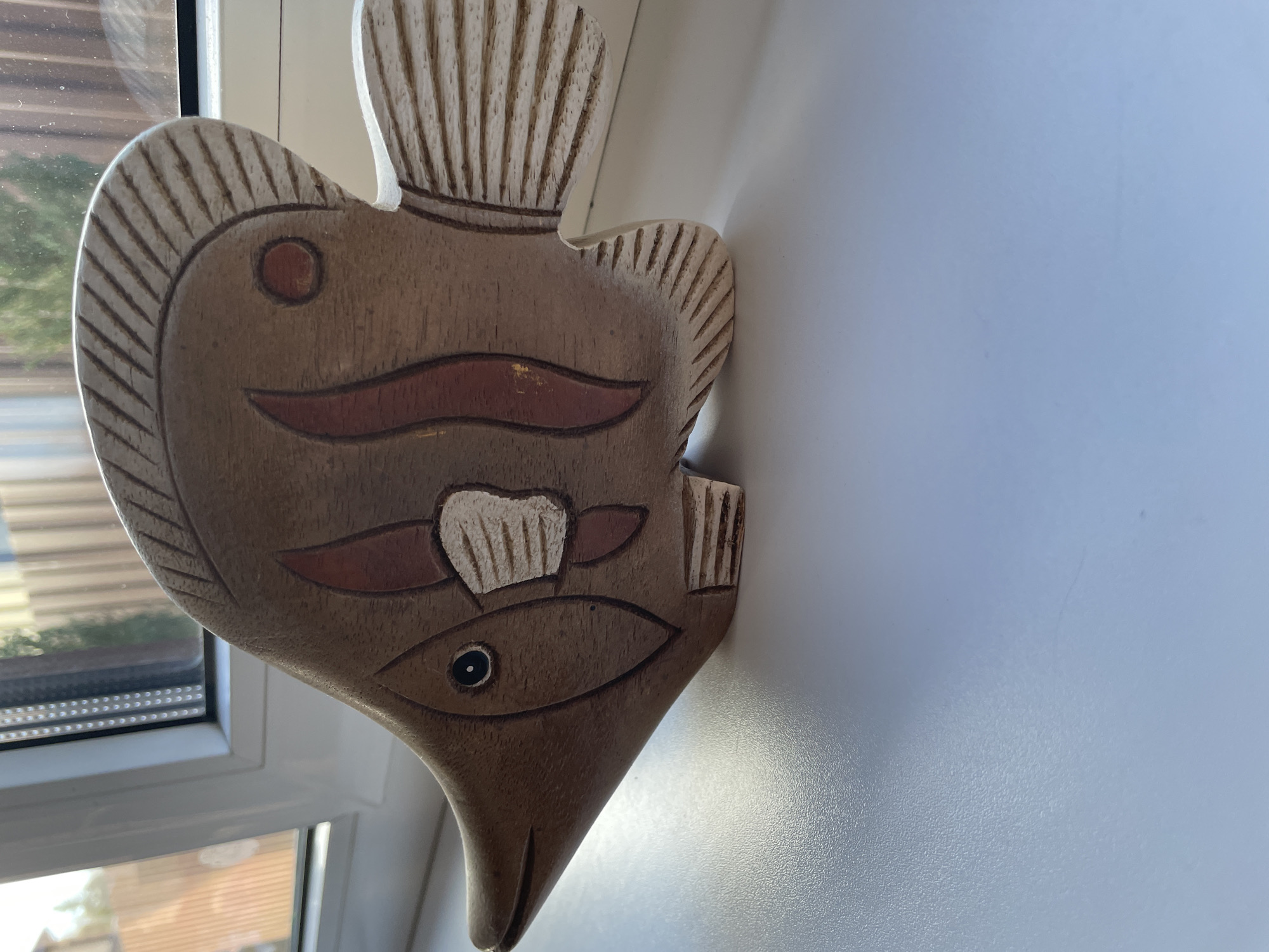 A wooden fish decoration is hanging on a white wall next to a window. The fish has carved details representing eyes, fins, and a tail. The background includes a window with visible outdoor scenery, suggesting an indoor location. The wall is clean and uniformly colored. The scene appears to be in a residential or domestic setting. There is no location in the city of Лесной Пейзаж Кп (шарапово, Russia.