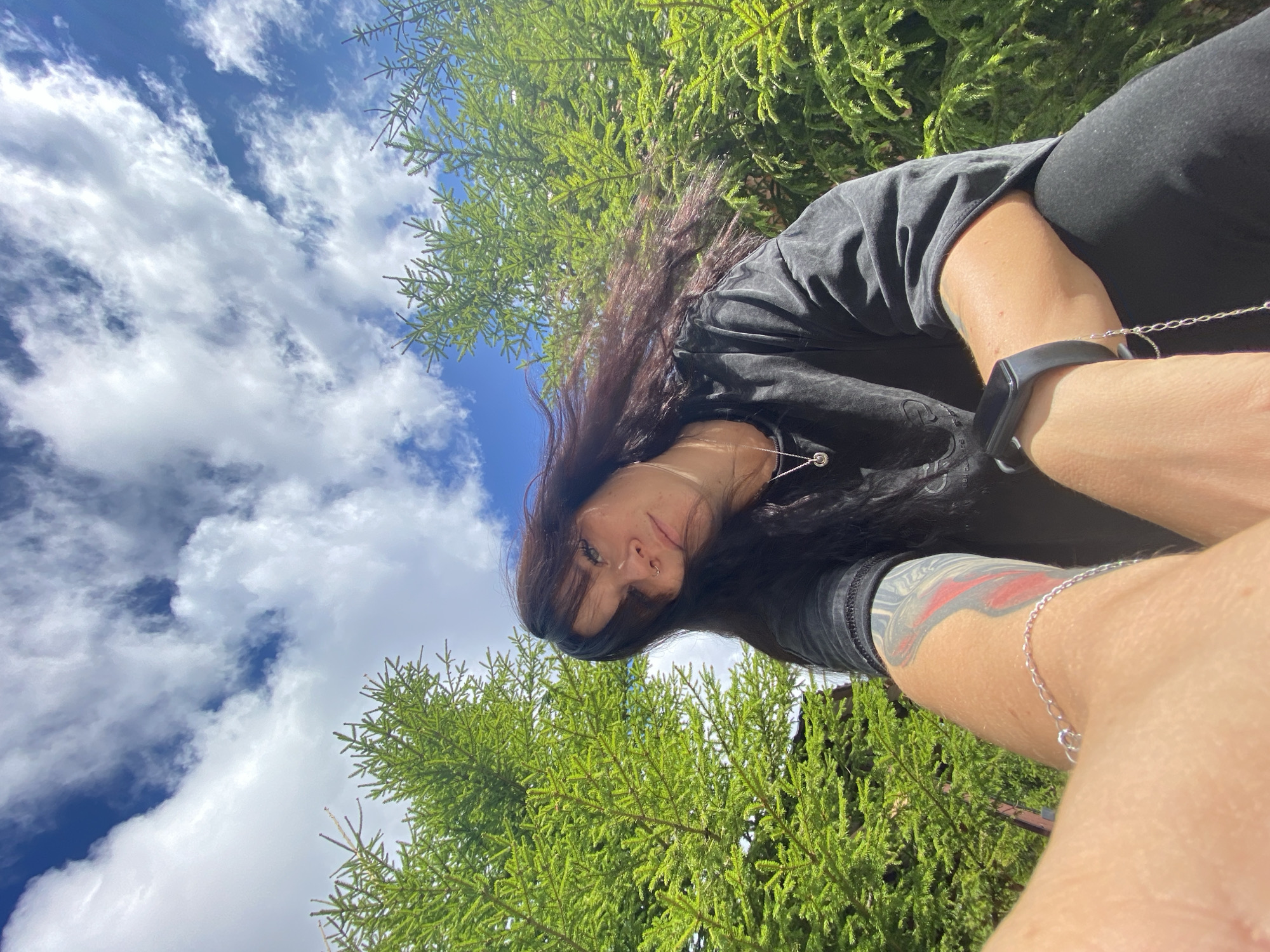 The image shows a woman with dark hair taking a selfie outdoors. She is wearing a dark t-shirt, black pants, and a black fitness tracker on her left wrist. A silver chain bracelet is also visible on her left wrist. She has a tattoo on her left arm. She is surrounded by green pine trees under a bright blue sky with scattered white clouds. The perspective is from below, looking up at the woman and the sky. No specific location within Krasnoflotskoe, Russia can be determined from the visual information provided.