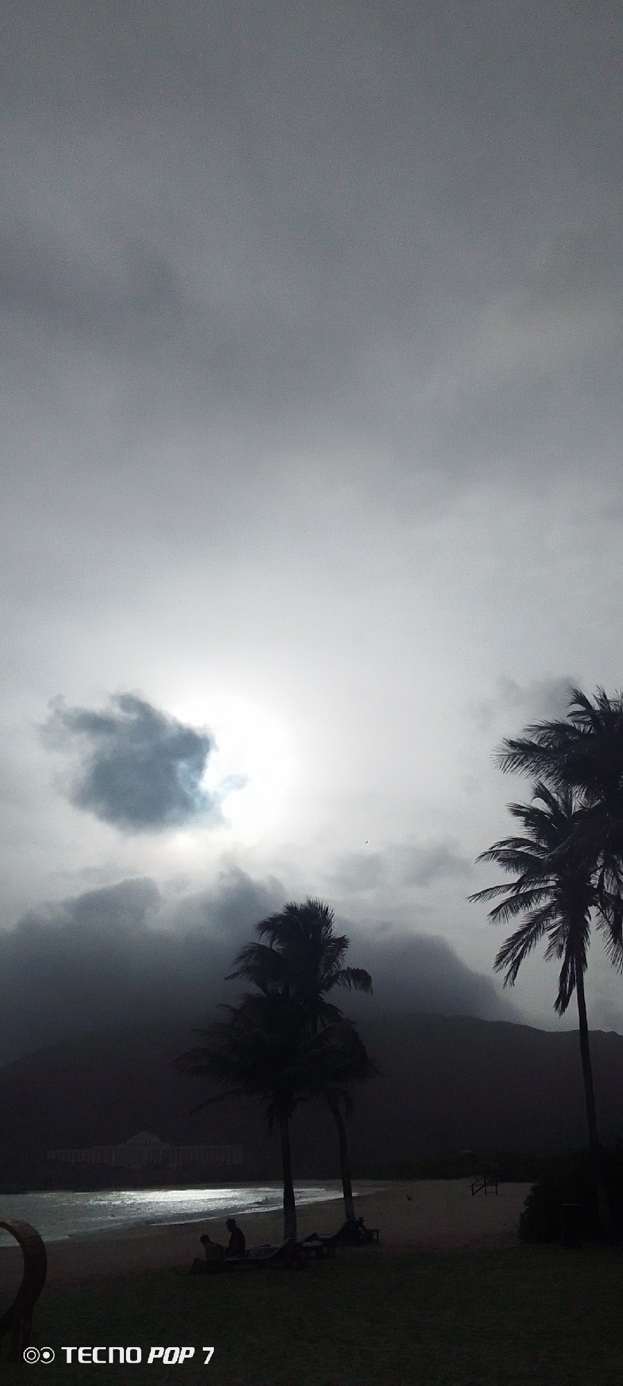 The image depicts a beach in Pedro González, Venezuela, under a cloudy, overcast sky. Two palm trees are visible in the foreground, with other trees further back near the beach. The sky is filled with grey clouds, obscuring the sun which is faintly visible. The beach is somewhat illuminated, with a couple of people sitting or lounging on the sand. Behind the beach, a dark hillside or mountain range is suggested. The bottom left corner shows a partial view of a light object. At the bottom, the phone watermark "TECNO POP 7" is visible.