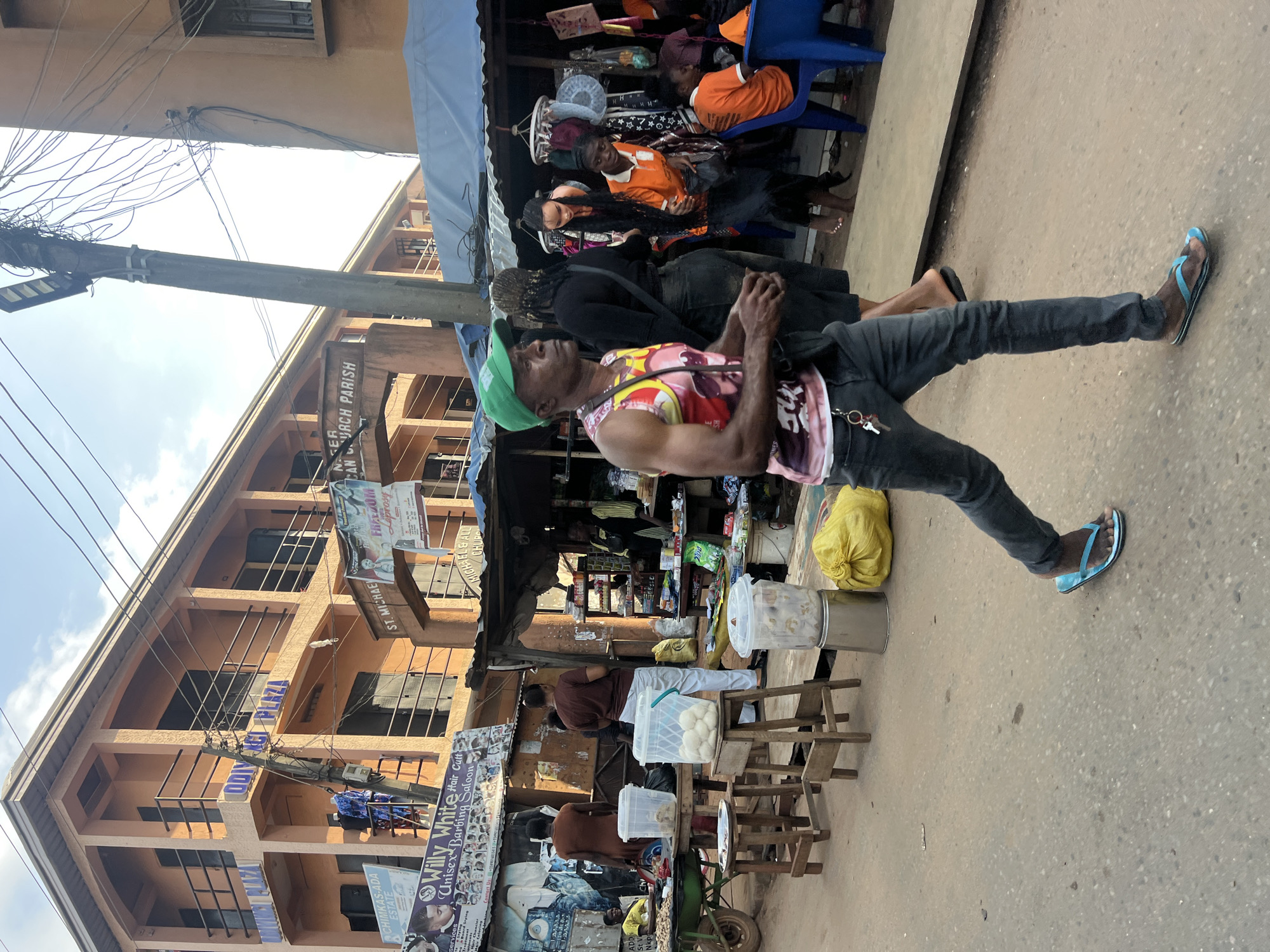 This is a vertical, eye-level shot of a bustling street scene in Nkpor, Nigeria. The foreground is dominated by a man walking towards the right, wearing a green cap, a sleeveless shirt with a colorful, abstract design, dark jeans, and blue flip-flops. He has his hands clasped in front of him and appears to be looking upwards or to the side.
Behind him, a market stall is set up, with various items displayed on shelves and tables. Several people are seen in and around the stall, including a woman in an orange shirt and dark skirt, and others in orange and black clothing. A mannequin with long black hair is also visible. The stall has a blue awning and is cluttered with goods.
To the left of the market, a multi-story building with an orange facade is visible. Signs are plastered on the building, including one that reads "ODIVIC PLAZA" and another indicating "ST. MICHAEL & ALL CHURCH PARISH". Another sign advertises "Willy White Hair Cult Unisex Barbing Saloon".
In the background, more buildings and overhead wires are visible, suggesting a dense urban environment. The sky is bright, indicating daytime, and the lighting suggests it might be a sunny day. The scene appears to be a typical commercial area in Nigeria, with vendors selling their wares and people going about their daily activities. The overall impression is one of lively commerce and community interaction.
