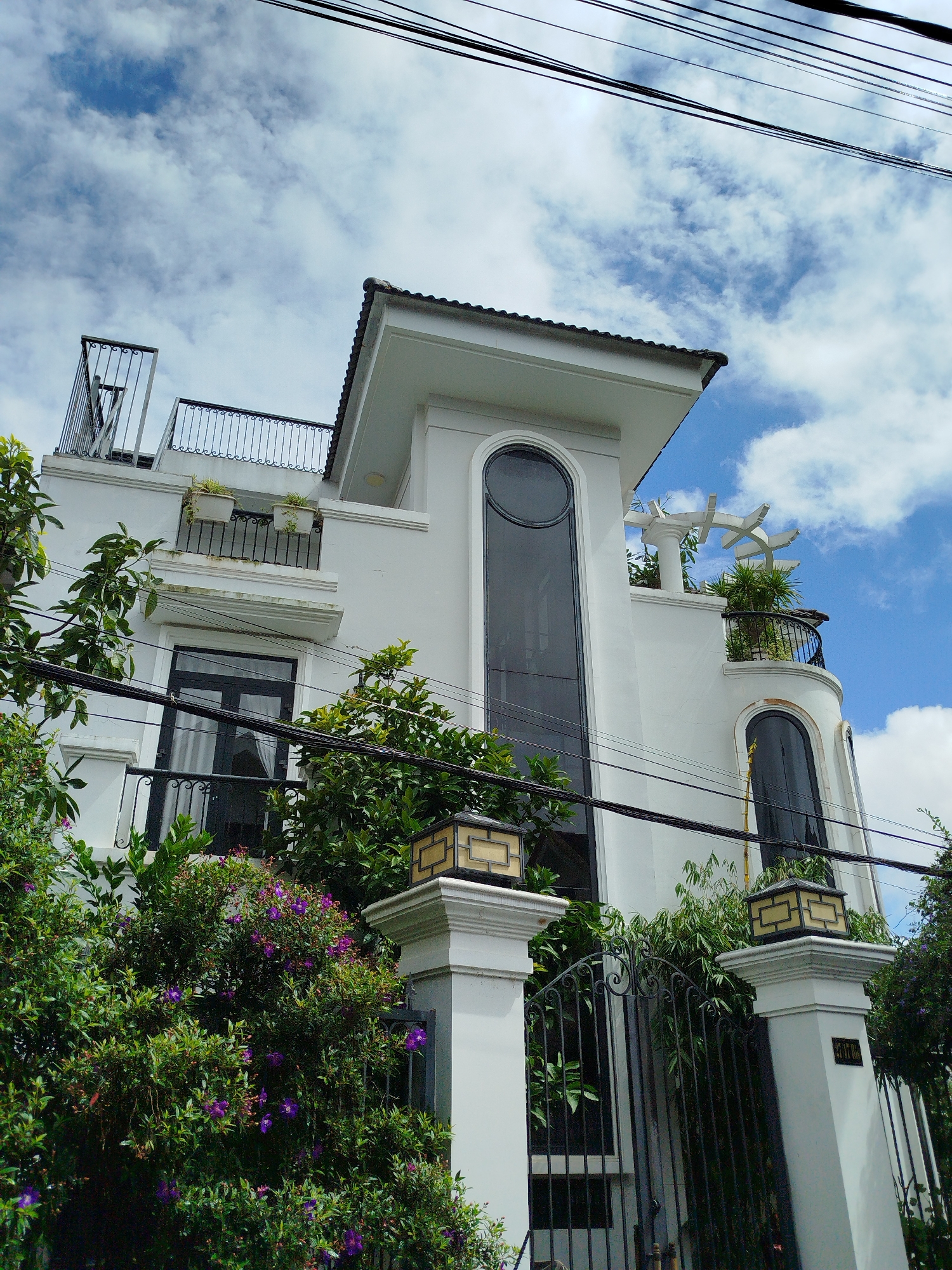 The image shows a modern white building with several balconies, a rooftop terrace with a pergola, and large, arched windows. The building is surrounded by lush green foliage and flowering purple plants. Overhead, power lines are visible against a partly cloudy blue sky. The facade of the building features decorative pillars and ornate metal railings on the balconies and gate. A street number plate is visible on the right-hand pillar. The scene appears to be a residential property in Da Lat, Vietnam, characterized by its tropical vegetation and architectural style.