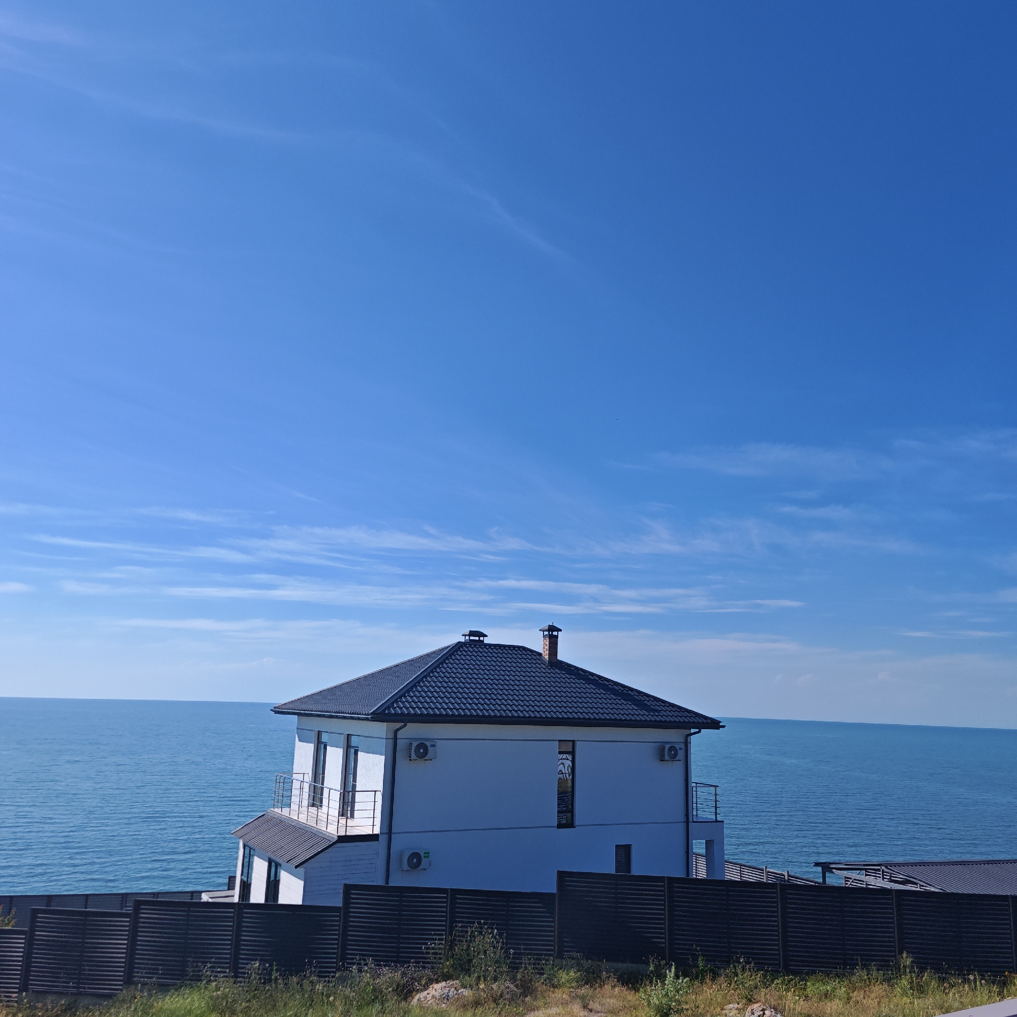 A modern white house with a dark tiled roof is situated on a grassy hill overlooking a calm blue sea. The house has multiple windows, balconies with railings, and two air conditioning units mounted on its exterior walls. A chimney rises from the roof. In front of the house, a dark horizontal slatted fence runs across the foreground, with dry grass and scattered rocks visible beneath it. The vast expanse of the bright blue sky, streaked with thin, wispy white clouds, dominates the upper portion of the image. The sea is a deep blue and appears placid, meeting the sky at the horizon. The sun is high, suggesting midday or early afternoon. The scene evokes a sense of tranquility and peacefulness, characteristic of a seaside dwelling on a clear, sunny day. There is no visible text in the image.