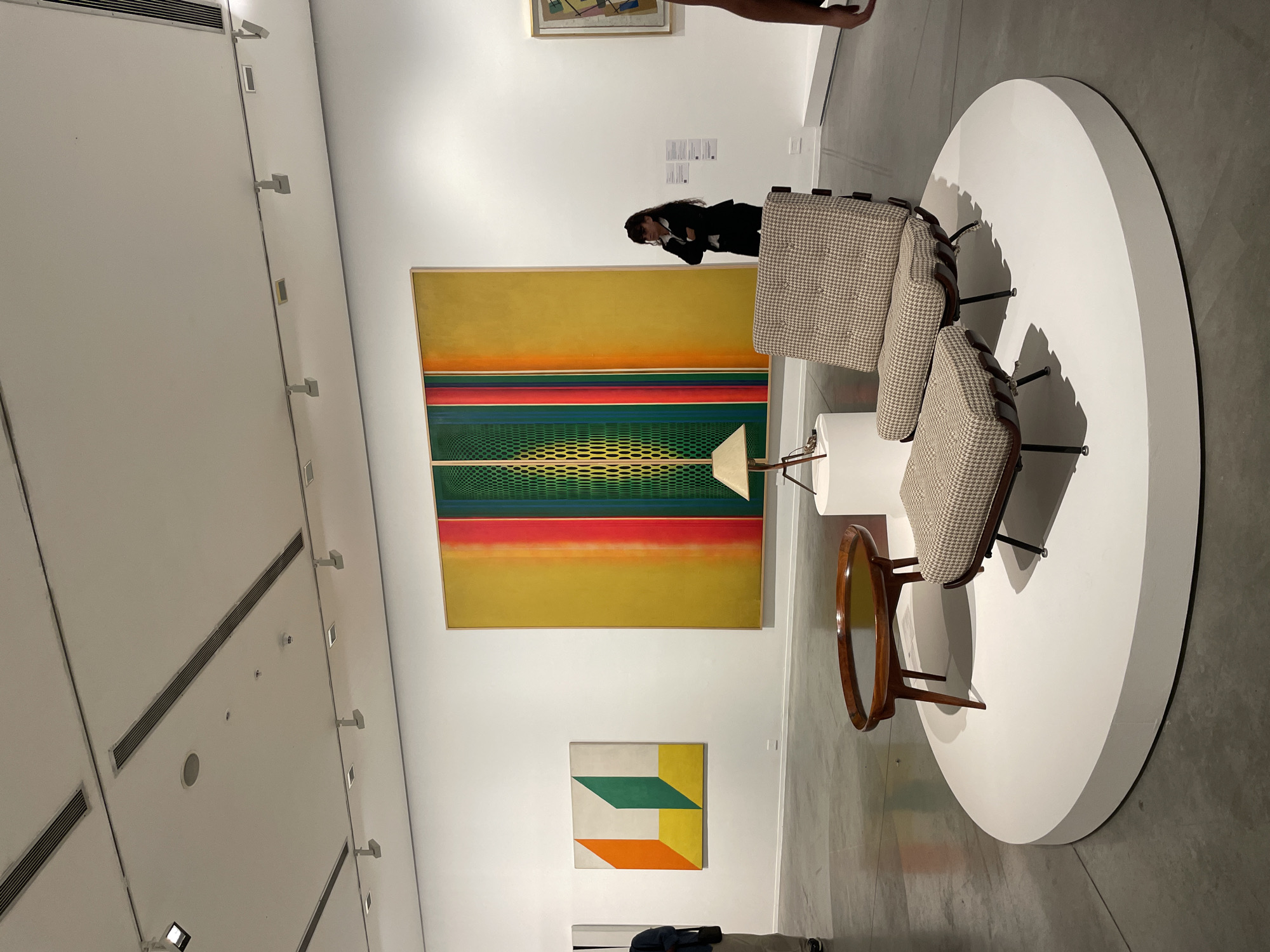 The image depicts an art gallery interior, likely in Buenos Aires, Argentina, though specific location cues are absent. The scene centers on two mid-century modern armchairs with patterned upholstery, positioned on a large, circular white platform. A small, round wooden table is placed between the chairs. Behind them hangs a large abstract painting with horizontal bands of yellow, red, and green over a grid pattern, alongside a smaller geometric painting. A woman, dressed in dark clothing, is standing near the paintings, appearing to be viewing or photographing the art. The gallery space has white walls and a minimalist aesthetic. Ventilation ducts and lighting fixtures are visible on the ceiling. An additional person is partially visible in the background. The overall atmosphere is one of quiet observation in a contemporary art setting.