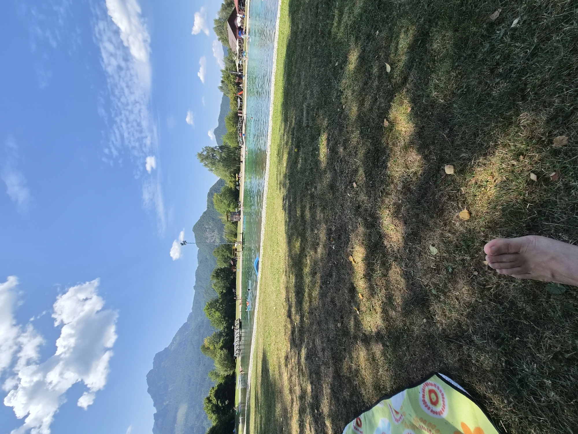 The image shows a sunny day at an outdoor swimming area in Landl, Austria. In the foreground, a person's bare foot and a patterned blanket lie on a grassy area. Beyond the grass, a large, clear pool of water is visible, with several people swimming and relaxing on inflatable toys. A wooden walkway extends into the pool. In the background, lush green trees and mountains form a scenic backdrop under a bright blue sky with fluffy white clouds. Buildings, including what appears to be a restaurant or service area, are situated along the edge of the water. A tall lighting pole is also visible. The overall impression is a recreational scene in a natural, mountainous setting.