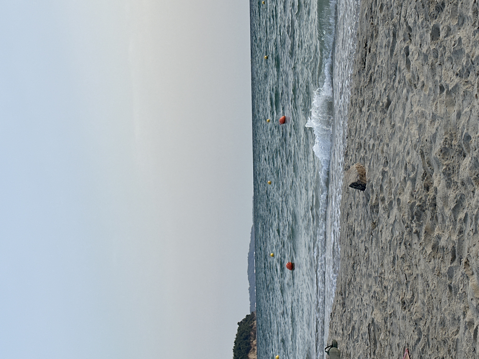 The image depicts a serene beach scene in Kassandra Municipal Unit, Greece, under a pale sky. The foreground is dominated by sandy beach textured with footprints. A single, rough-hewn rock rests on the sand. To the left, partially visible on the sand, are what appear to be a light-colored bag and a pair of flip-flops.
The middle ground features the gentle lapping of waves against the shore. Beyond the immediate surf, the sea is a mottled blue-green, exhibiting a moderate chop. Several buoys are visible on the water's surface: a prominent orange one in the center-left, and several smaller yellow ones scattered further out. A wave is captured in mid-break, producing white foam.
In the background, a lush green, tree-covered headland juts out from the left, creating a natural cove. Further in the distance, a hazy, undulating shoreline is visible under the pale sky, suggesting more landmass or islands. The overall atmosphere is one of tranquility and natural beauty.