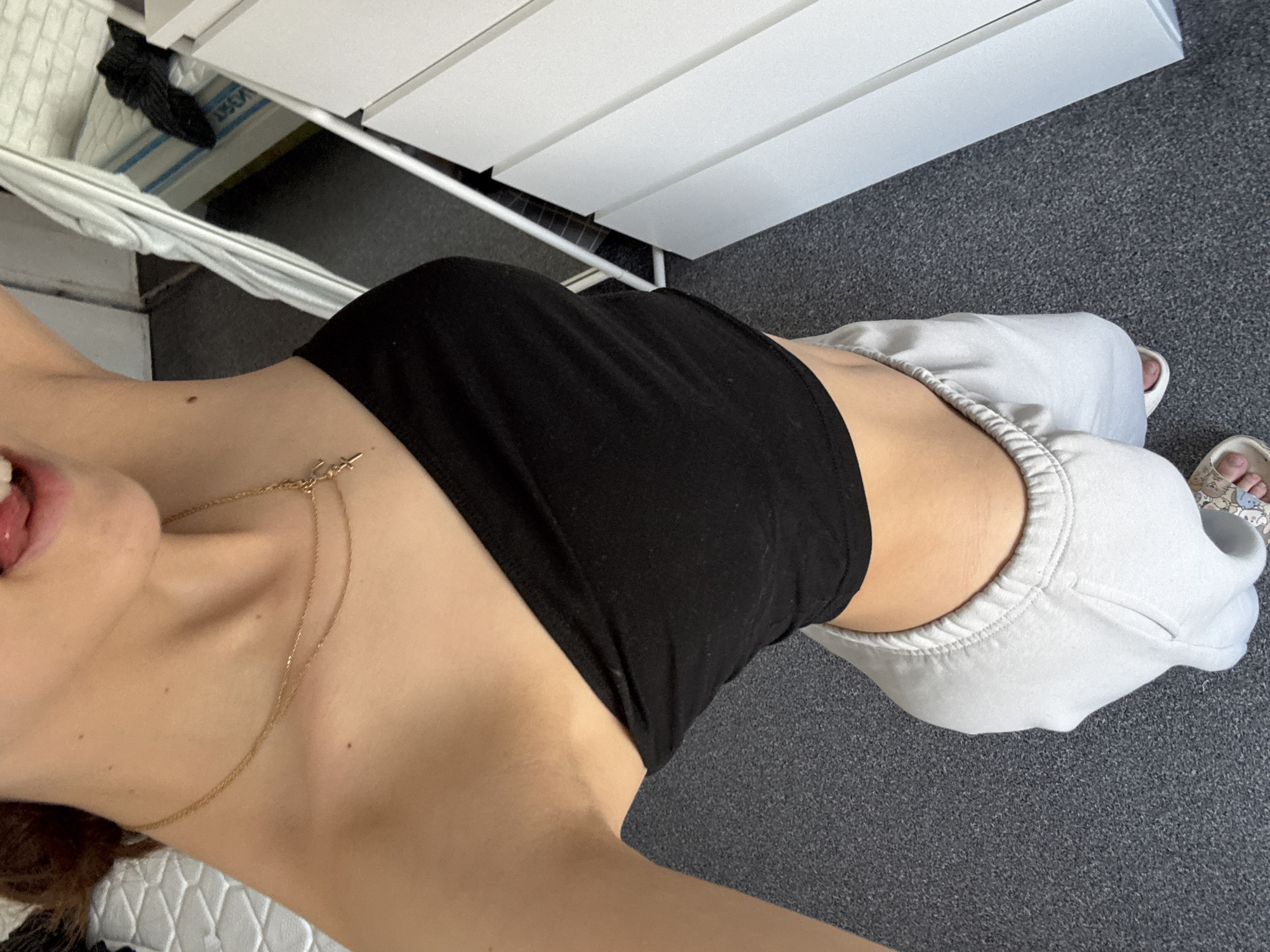 A view from above shows a person wearing a black strapless top and light grey sweatpants standing on a grey carpeted floor. The person is also wearing gold necklaces. The location is likely inside a room with white drawers and what appears to be a clothes drying rack. The location in Sofia, Bulgaria cannot be determined from the image.
