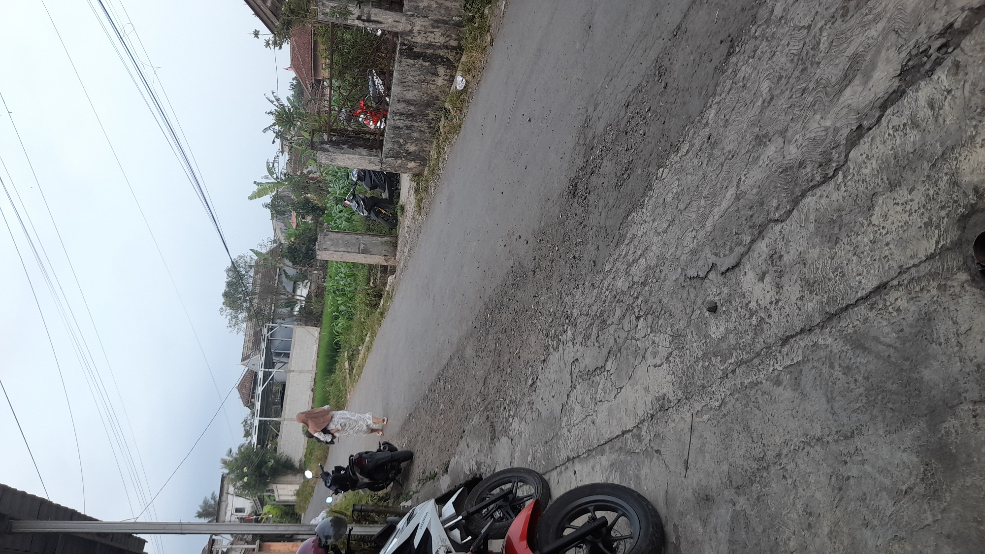 The image depicts a street scene in Selaawi, Indonesia, under an overcast sky. The foreground features the pavement, showing signs of wear and tear with cracks and textured patterns. To the left, several motorcycles are parked on the paved area. One person, a woman wearing a headscarf and a long dress, is walking away from the camera on the asphalt road. The road extends into the middle ground, where more motorcycles are visible parked near a wall and gate. Beyond the road, there is a field of green crops, likely corn, adjacent to buildings. The background reveals a residential area with houses and trees, characteristic of a village or town. The overall impression is a quiet, everyday moment in a semi-urban or rural Indonesian setting.