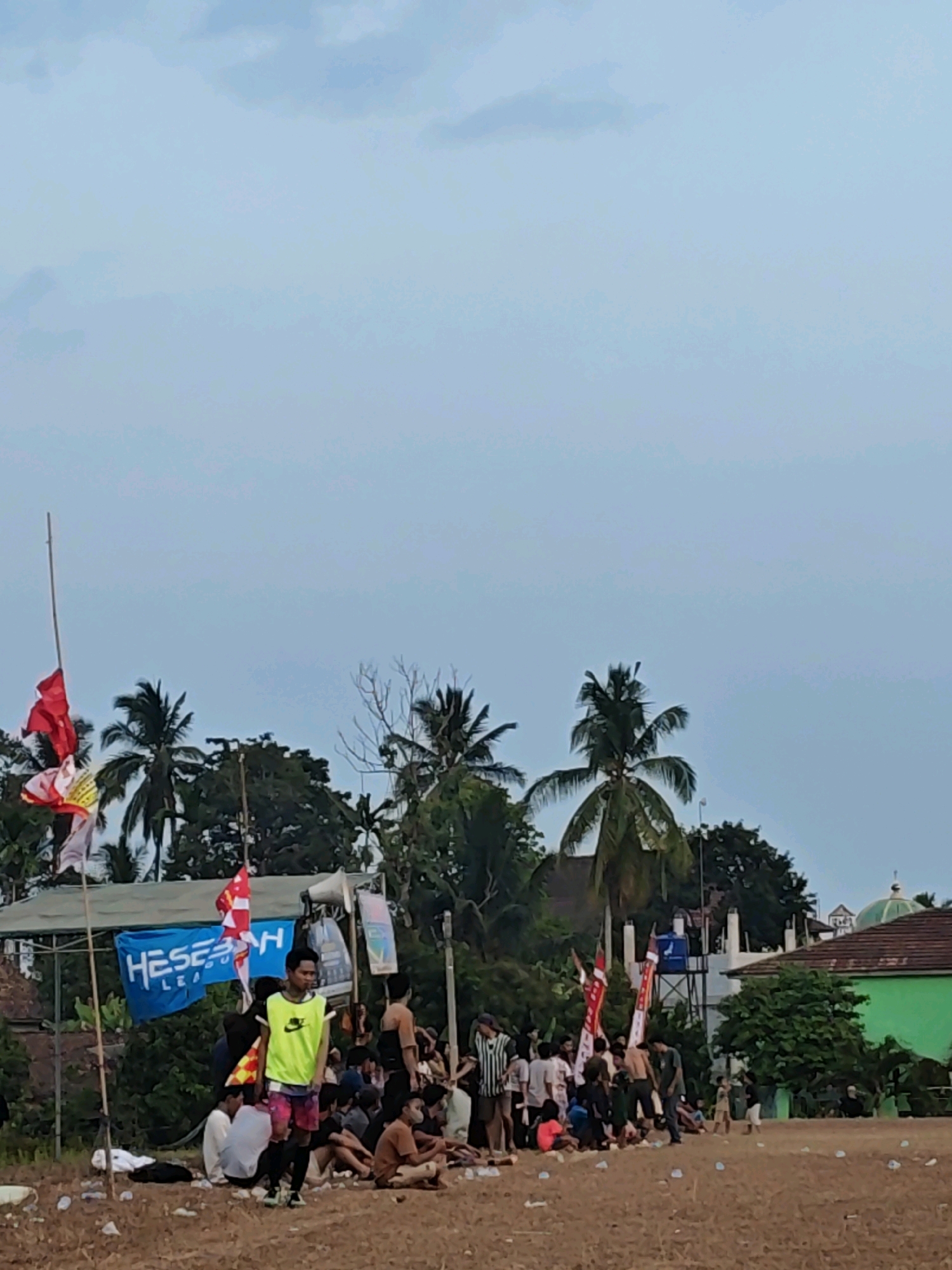 The image depicts an outdoor scene, likely in Karang Sari, Indonesia, with a gathering of people on a dirt field. The sky is overcast with a pale blue hue. In the foreground, the ground is covered in dry, brown dirt with scattered plastic debris. A group of individuals, varying in age and gender, are visible, some sitting on the ground and others standing. One young man in the center wears a bright yellow vest over a t-shirt and patterned shorts, standing and looking towards the right. Behind him, a blue banner with white lettering, possibly "HESEKH LEAGUE," is displayed. Red and white flags are also present, some on poles. Palm trees and other foliage are scattered throughout the scene, with a building and a green domed structure visible in the background. The overall atmosphere suggests a casual event or gathering, possibly a sporting event given the flags and banners.