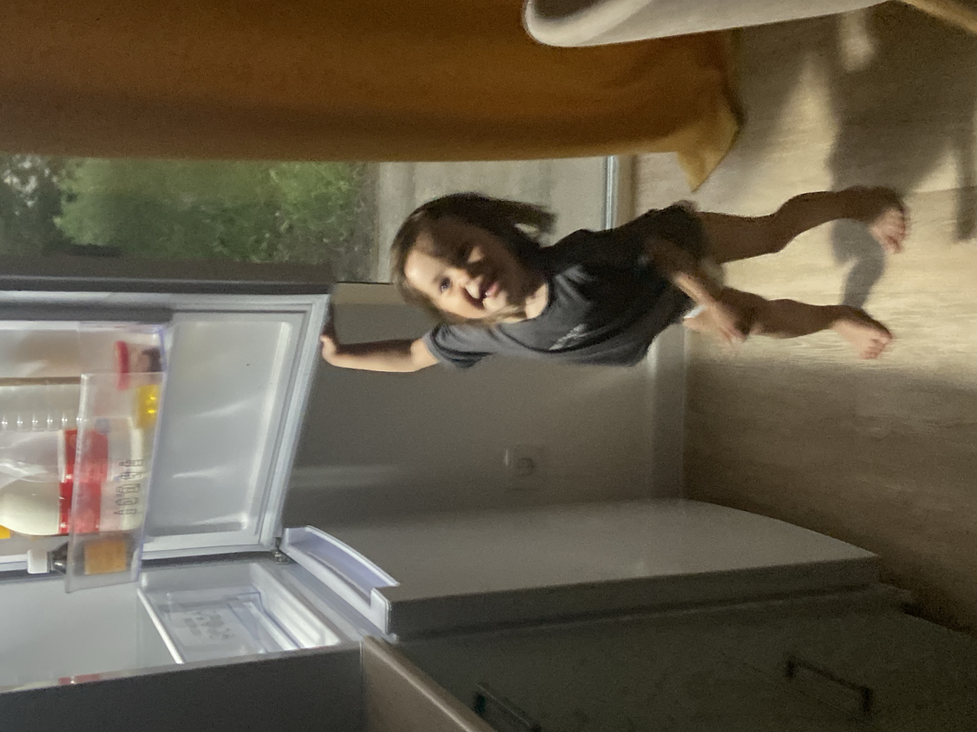A young child opens a refrigerator in a room. The child has short brown hair and wears a gray shirt. The refrigerator is white and contains food items. Outside the refrigerator, a window shows green foliage. The room also contains a light-colored chair or sofa with a brown cushion. The child is smiling. No information indicates this image was taken in Tyumen, Russia.