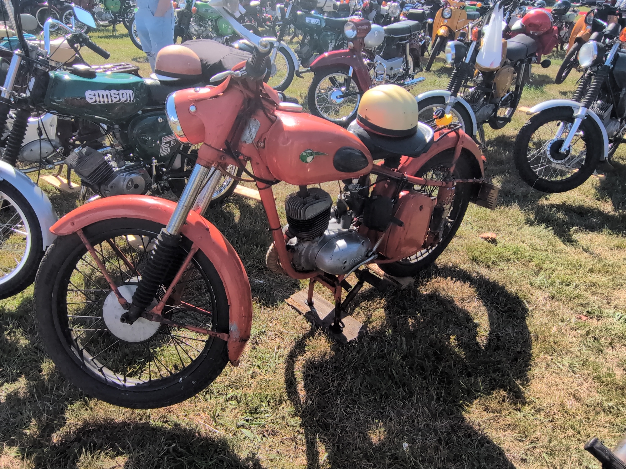 The image shows a gathering of vintage motorcycles parked on a grassy area, possibly during an outdoor event. In the foreground, a prominent red motorcycle with a yellow helmet on its seat is angled towards the viewer. This motorcycle features a spoked wheel, a visible engine, and a fender. Behind it, a green motorcycle with "SIMSON" branding is partially visible. Other motorcycles, some with yellow and red accents, are scattered throughout the background, suggesting a collection of classic vehicles. The ground is covered in dry grass, and shadows are cast by the motorcycles and possibly a person. The overall scene suggests a casual outdoor exhibition or meetup of motorcycle enthusiasts. The provided context indicates the location is the city of Hohenferchesar, Germany.