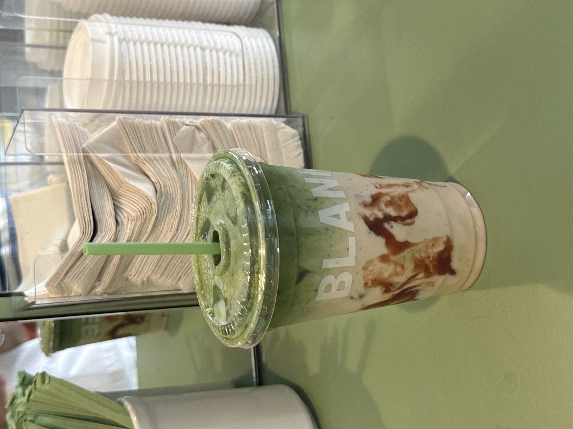 The image shows a partially filled plastic cup with a green straw, likely a matcha latte with brown caramel swirl, sitting on a light green surface. The cup is labeled "BLANK" in white lettering. To the left of the cup, there is a clear plastic dispenser containing stacks of paper napkins and white cups. In the bottom left corner, there is a container of green paper straws. The setting appears to be a cafe or restaurant, potentially in Boston, USA.