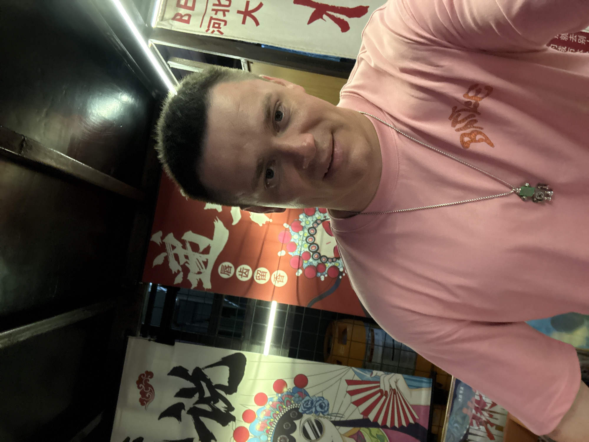 A man is taking a selfie in front of banners featuring Chinese opera imagery. He is wearing a pink t-shirt with an orange graphic and a silver chain with a green pendant. The background shows traditional Chinese artwork on banners. The scene is indoors, possibly a restaurant or market stall. There are no visible signs indicating the specific location within Qinhuangdao, China.